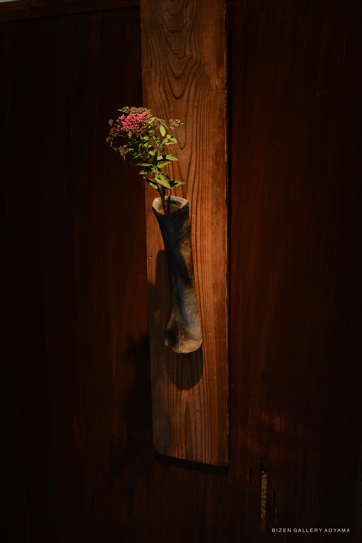 A ceramic vase with a flower arrangement, mounted on a wooden wall panel, showcasing traditional Bizen pottery craftsmanship.