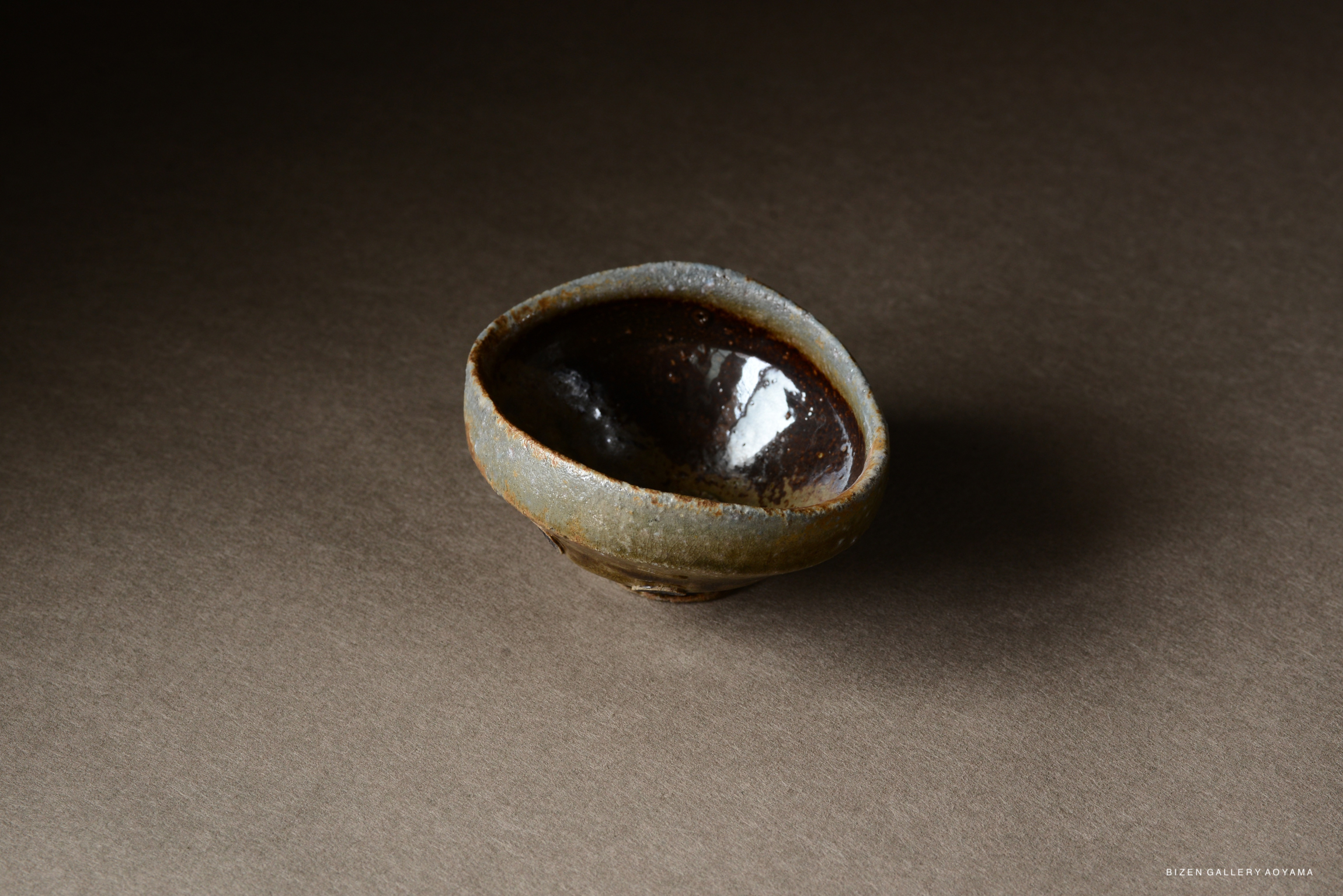 A small, uniquely shaped pottery bowl with a dark interior and a textured, light-colored exterior, placed on a neutral brown background.