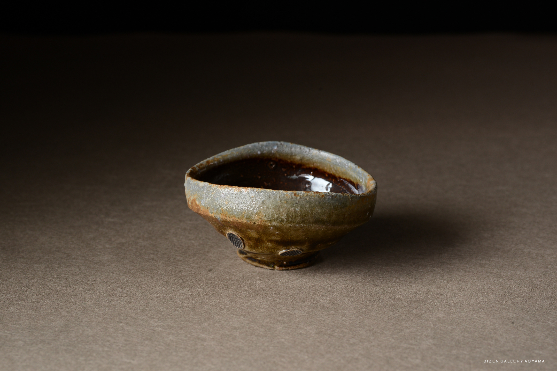 A small, rustic ceramic bowl with a unique shape and a glossy brown interior, set against a neutral background.