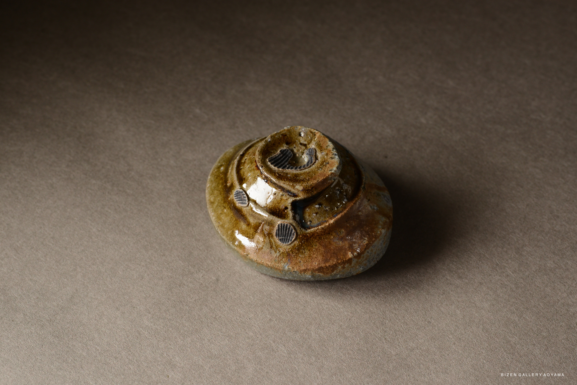 A close-up of a unique, textured ceramic piece with a glossy, earthy glaze, presented on a neutral background.