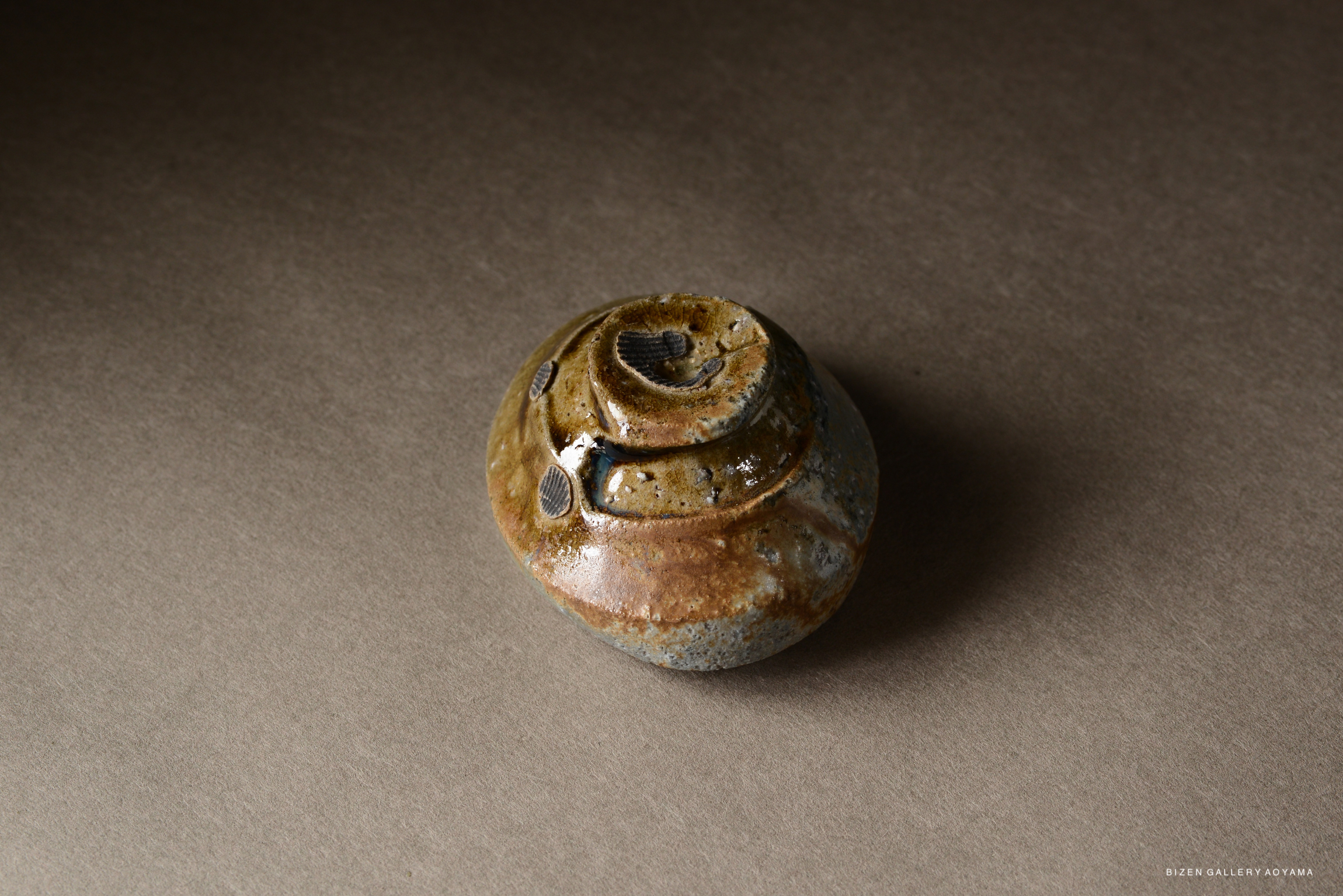A small, round, handcrafted pottery piece with a glossy brown and gray glaze, showcasing unique texture and design.