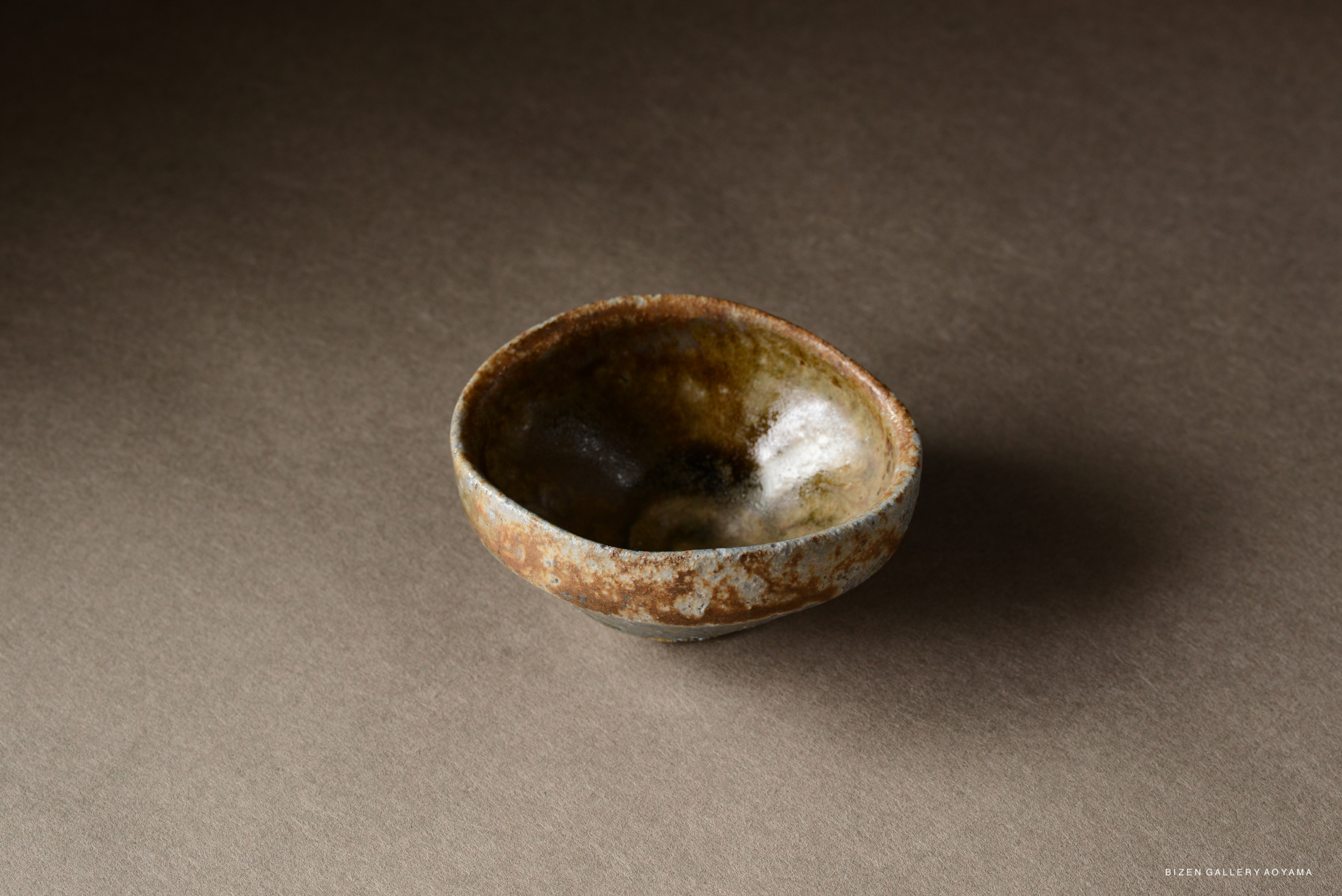 A small, rustic bowl made of pottery, featuring a mix of brown and gray glazes, placed on a neutral gray background.