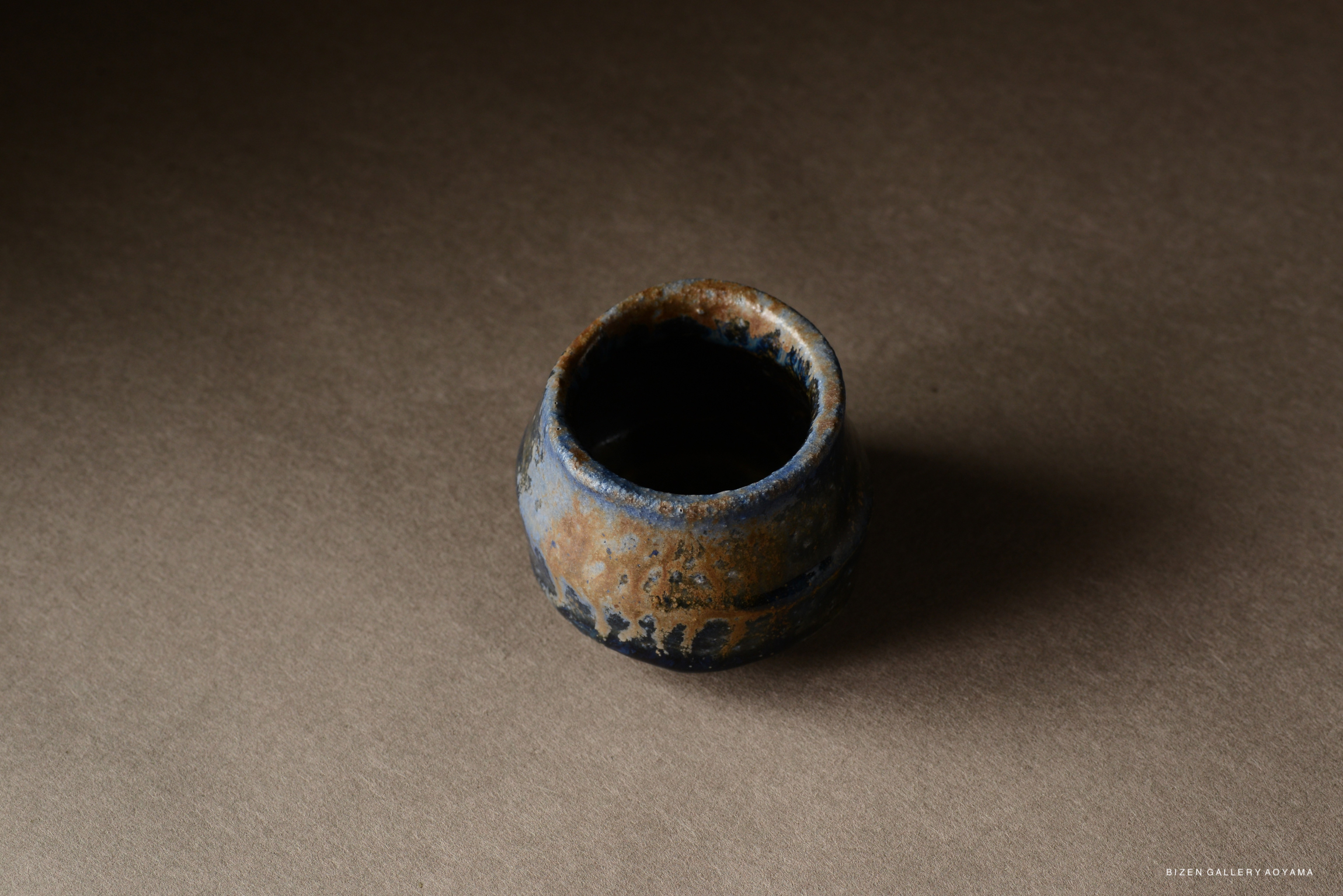 A small ceramic pot with a textured surface and a mix of blue and earthy tones, set against a neutral background.