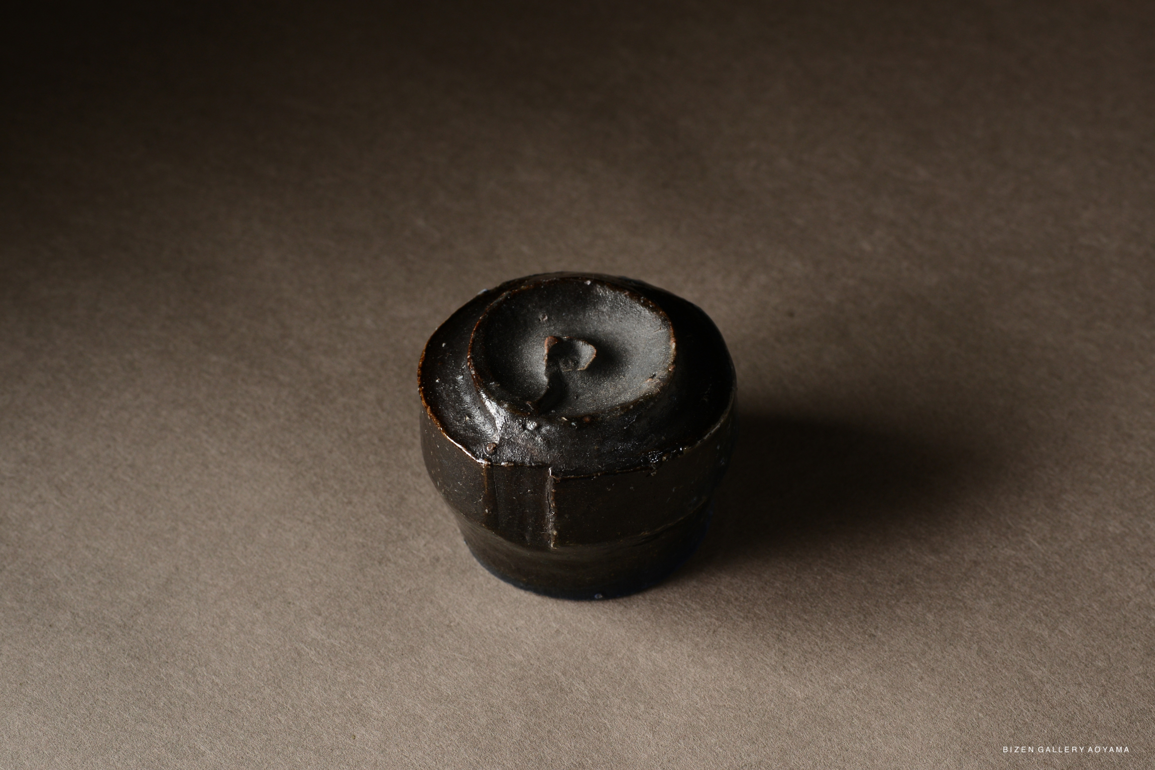 A close-up view of a small, dark ceramic vessel with a circular design on top, set against a gray background.