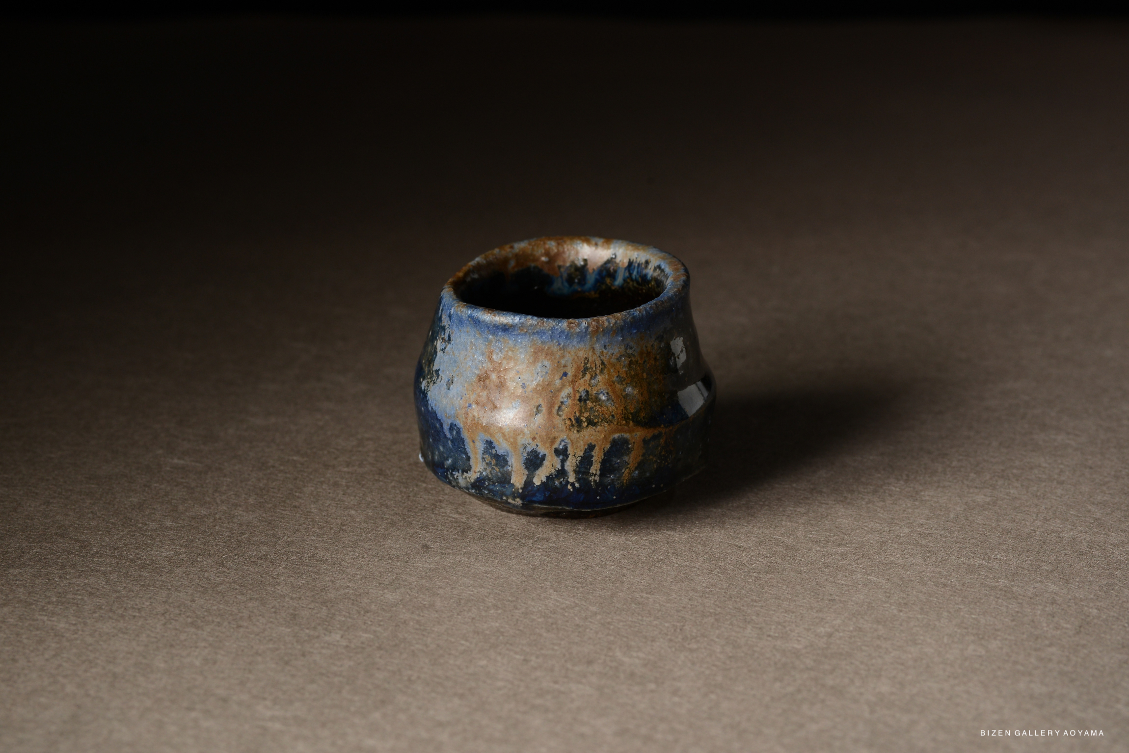A small, round pottery cup with a textured surface featuring blue and brown glazes, placed on a neutral background.