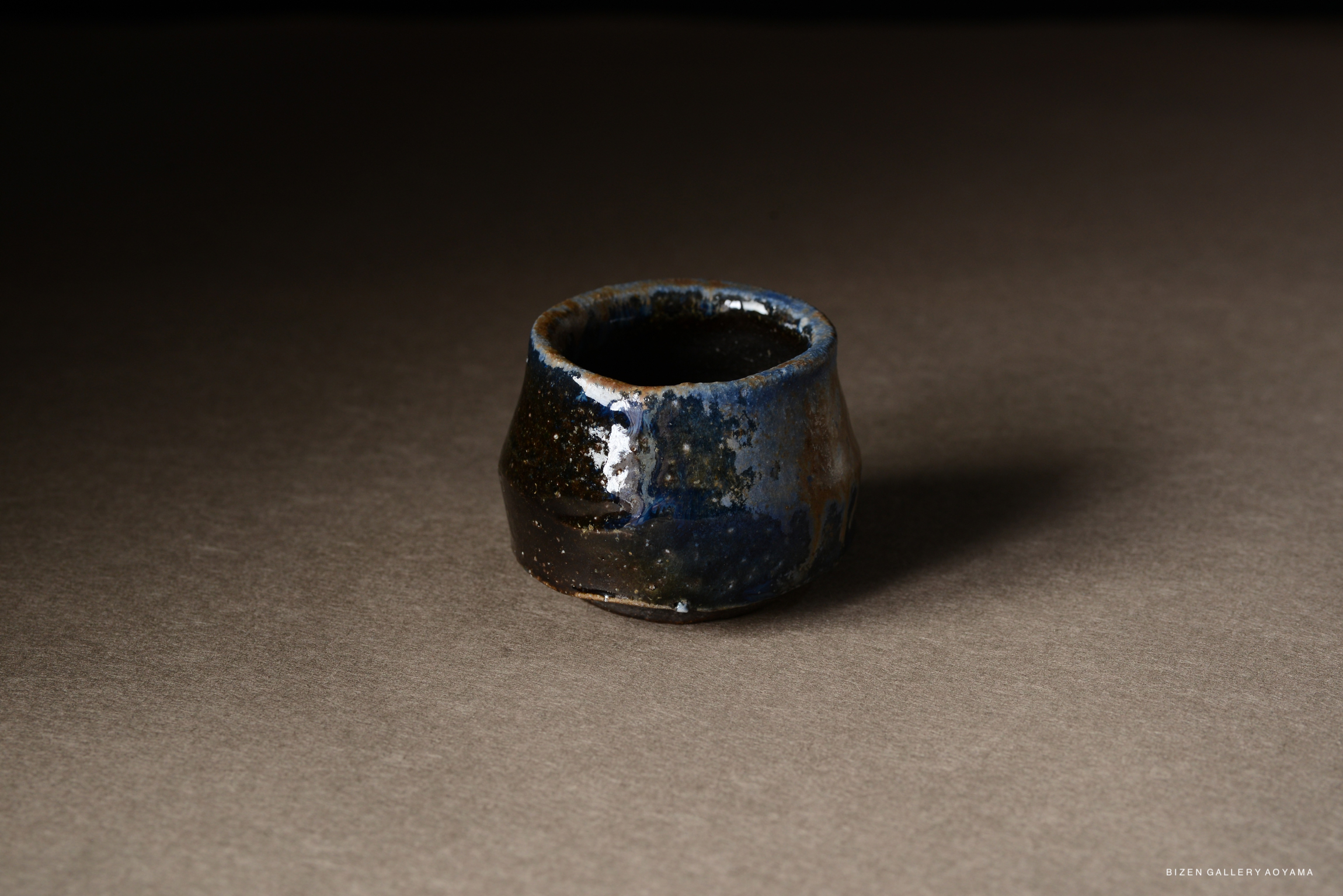 A small, handcrafted ceramic cup with a textured, dark blue glaze and a rustic finish, sitting on a neutral backdrop.