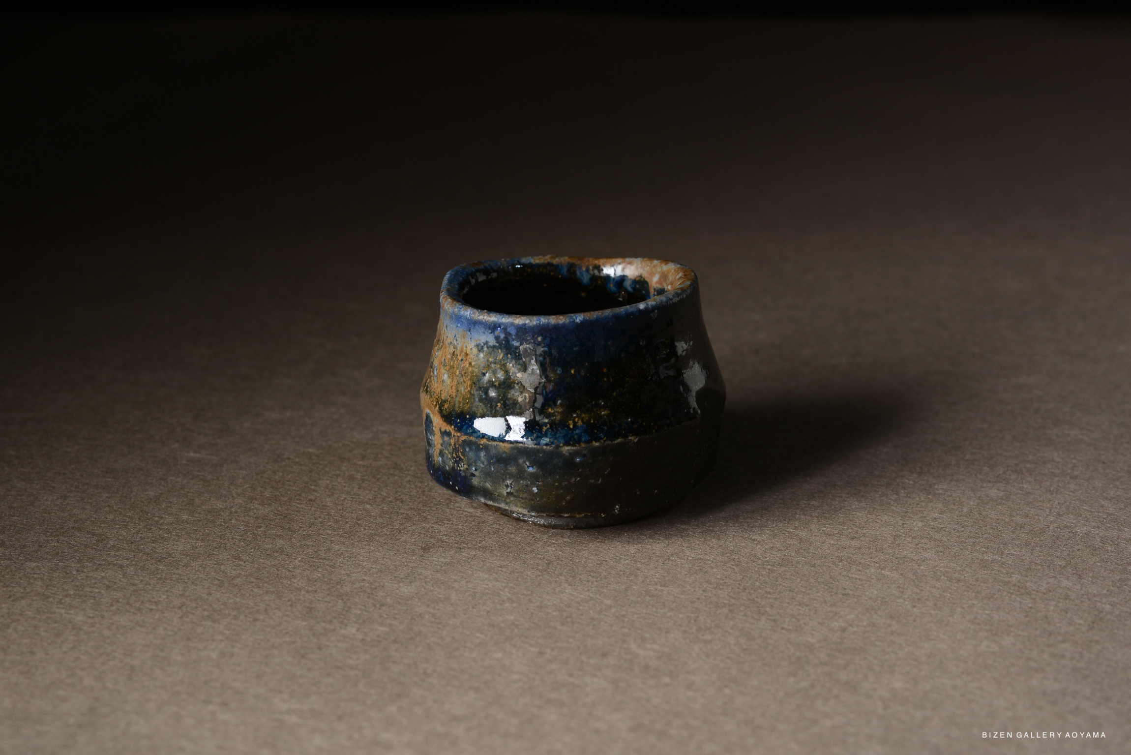 A close-up image of a uniquely shaped ceramic cup with a glossy blue and earthy glaze, set against a dark background.