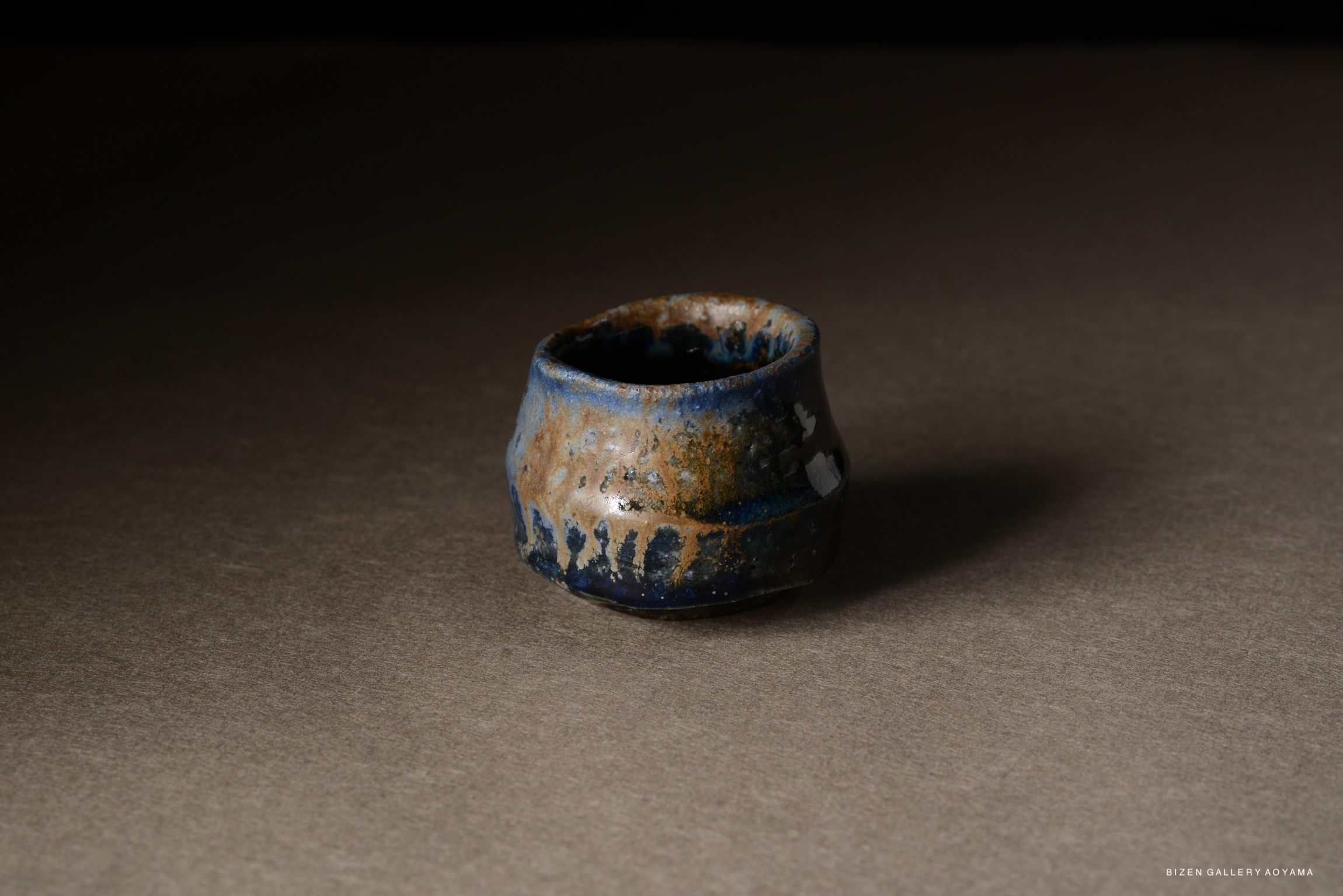 A small, handcrafted ceramic cup with a textured surface and a mix of blue and earthy tones, placed on a gray background.