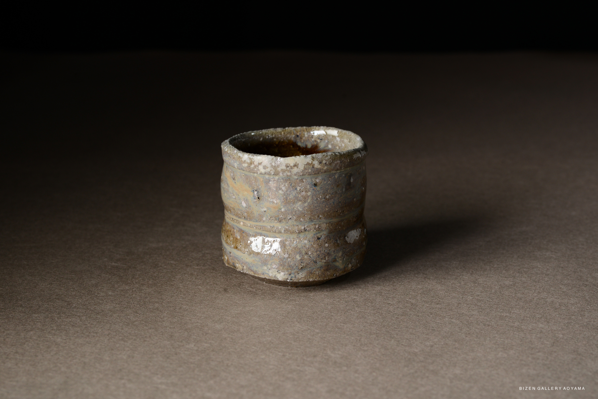 A small, rustic Bizen pottery vessel with a textured surface and muted colors, sitting on a brown backdrop.