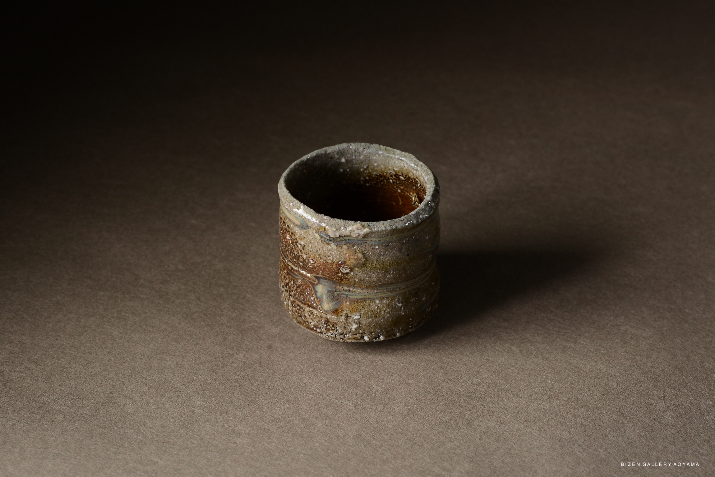 A close-up of a rustic Bizen pottery cup, showcasing its textured surface and earthy colors against a dark background.