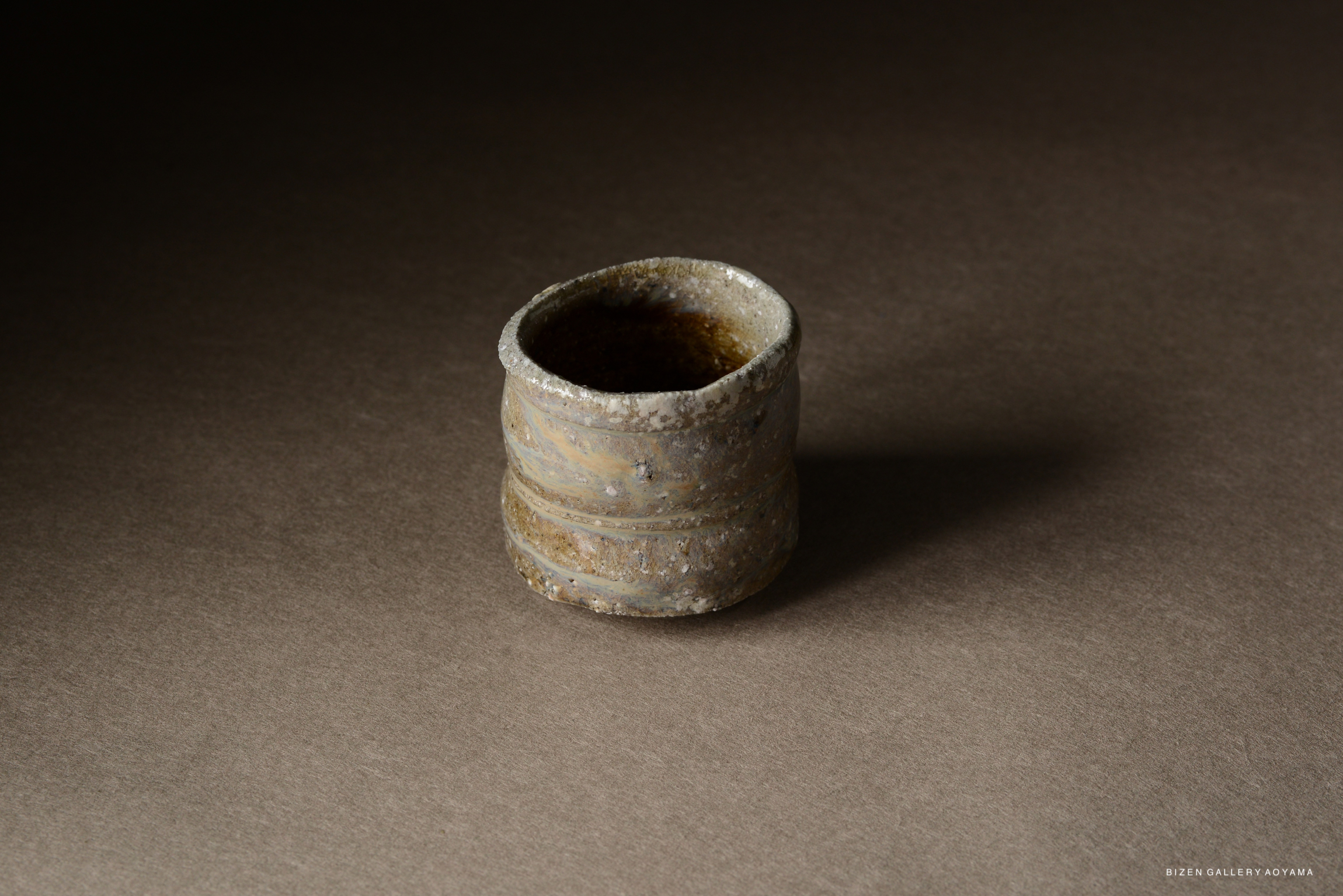 A textured ceramic cup with muted earthy tones displayed on a brown surface.