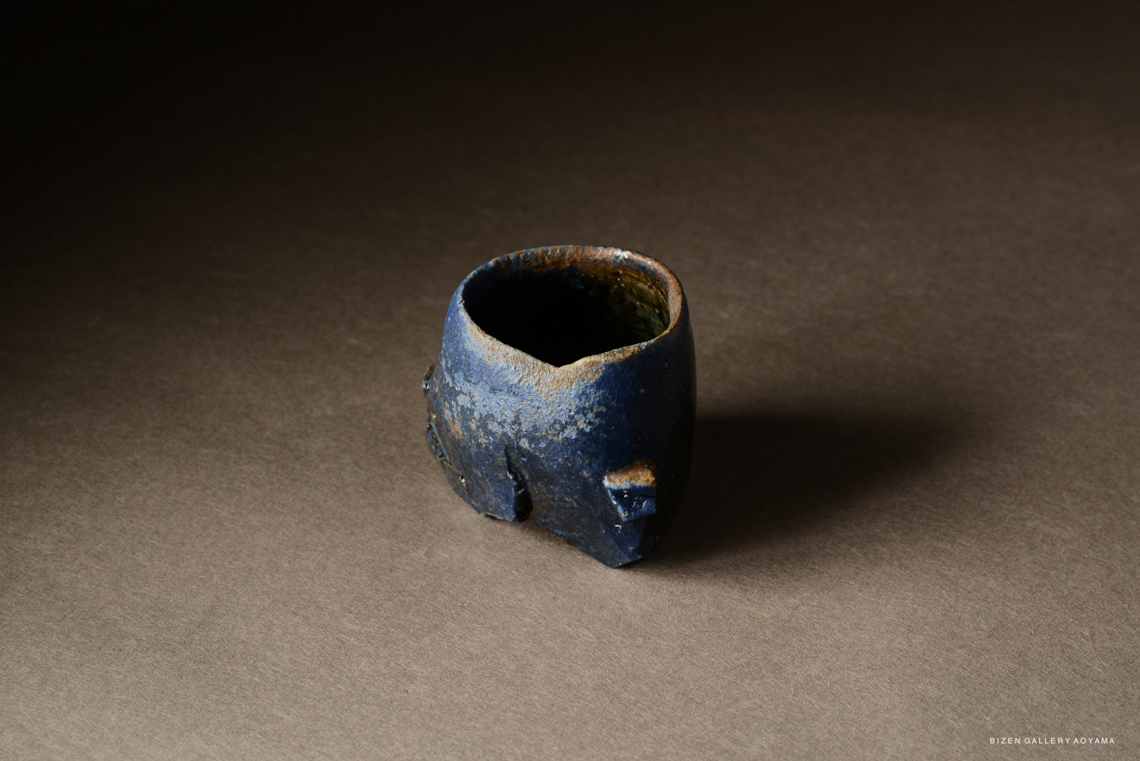 A uniquely shaped blue ceramic cup with a textured surface, positioned on a dark background.