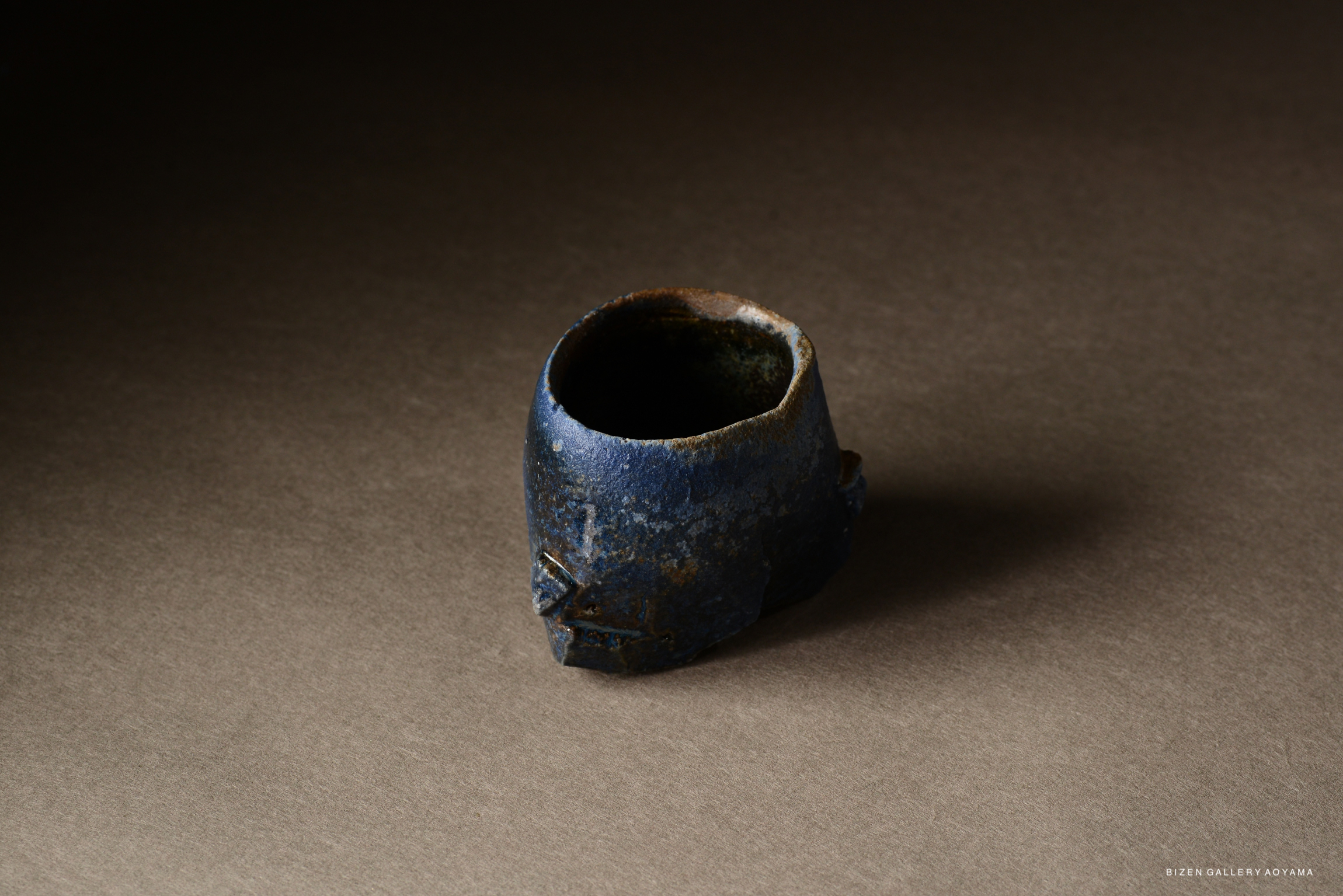 A close-up image of a unique Bizen pottery tea bowl featuring a textured blue exterior and a rustic, organic shape, set against a dark, neutral background.