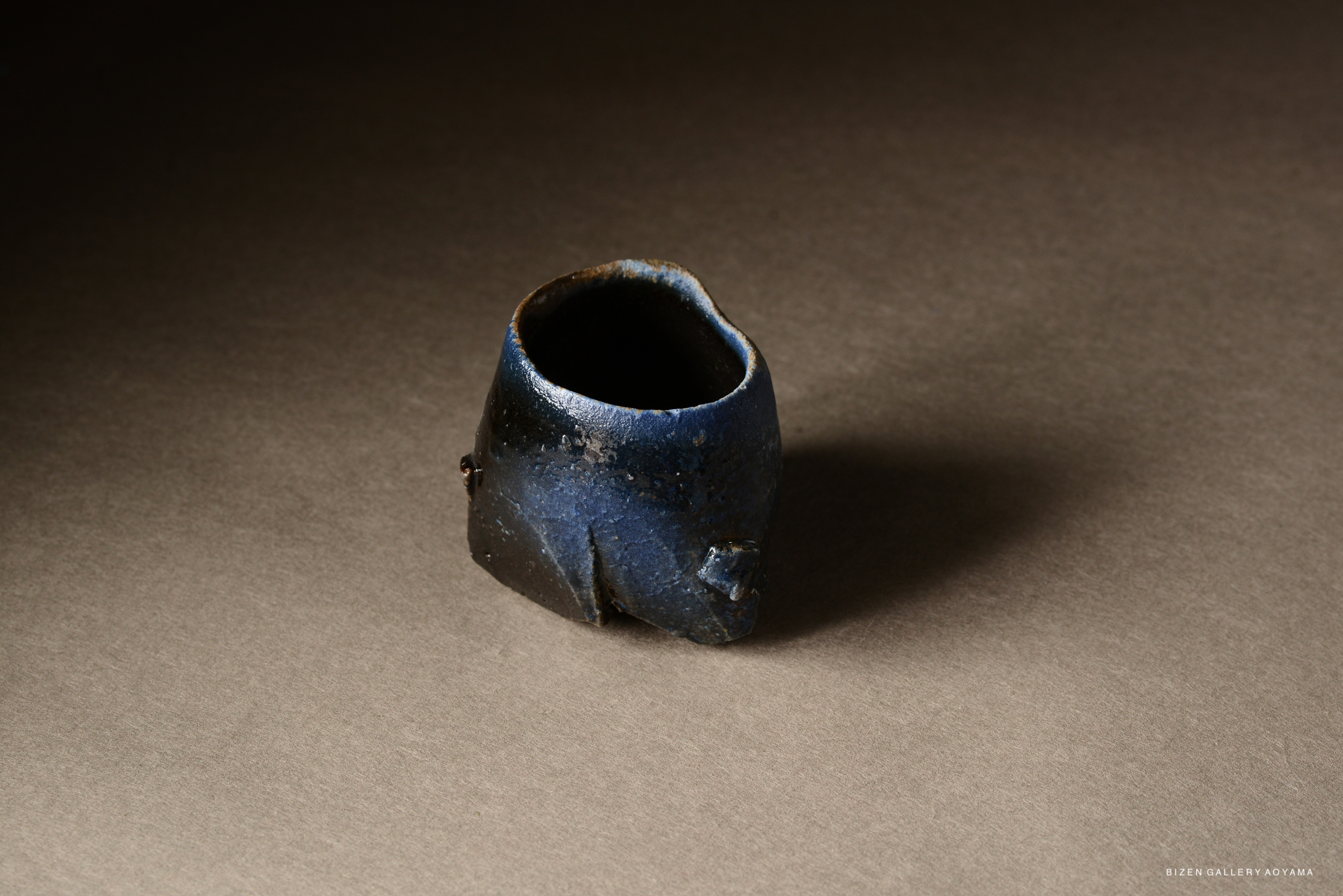 A unique blue ceramic vessel with an irregular shape, positioned against a neutral background.