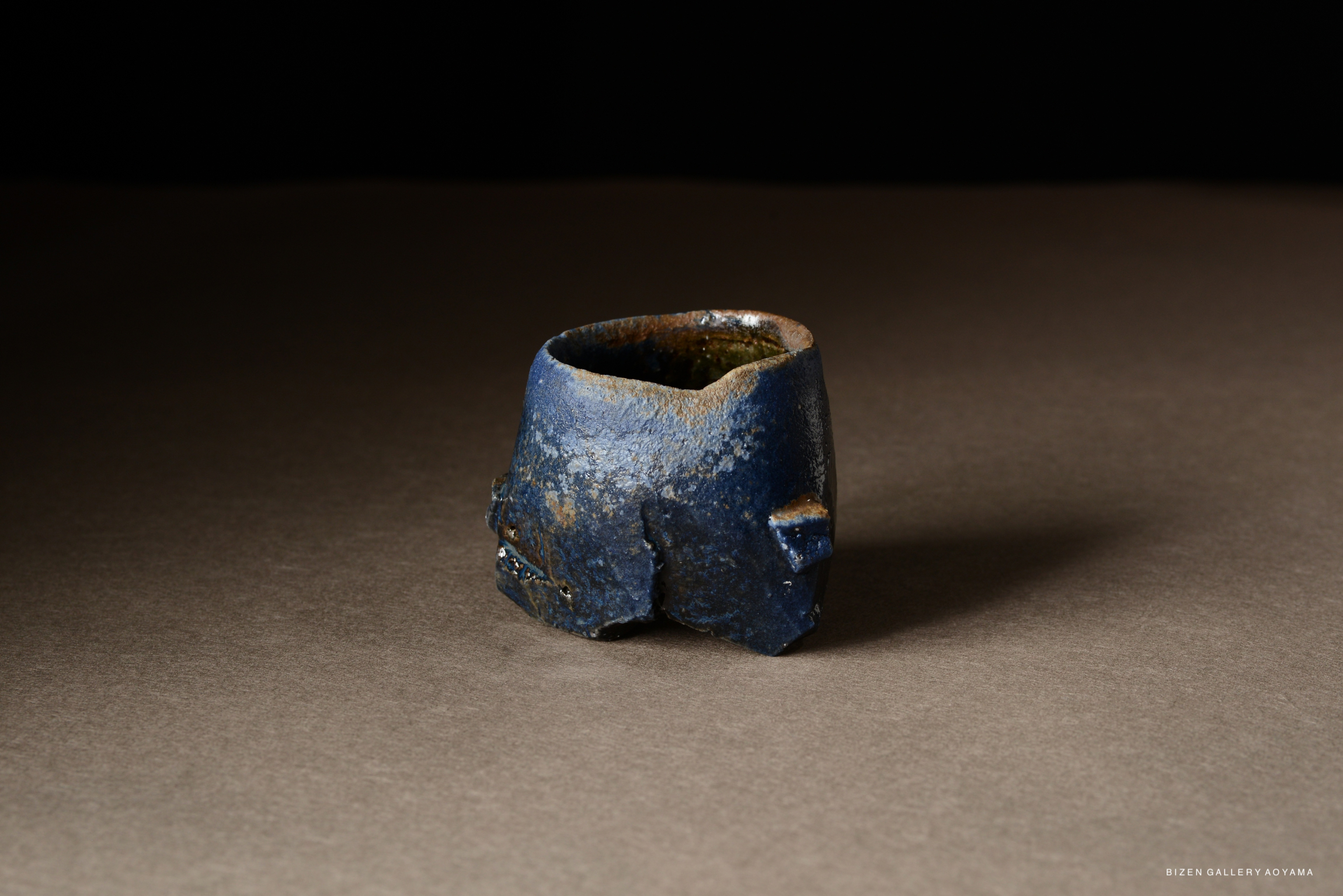 A handcrafted blue ceramic cup with a rustic finish, featuring uneven edges and a textured surface, placed on a neutral background.
