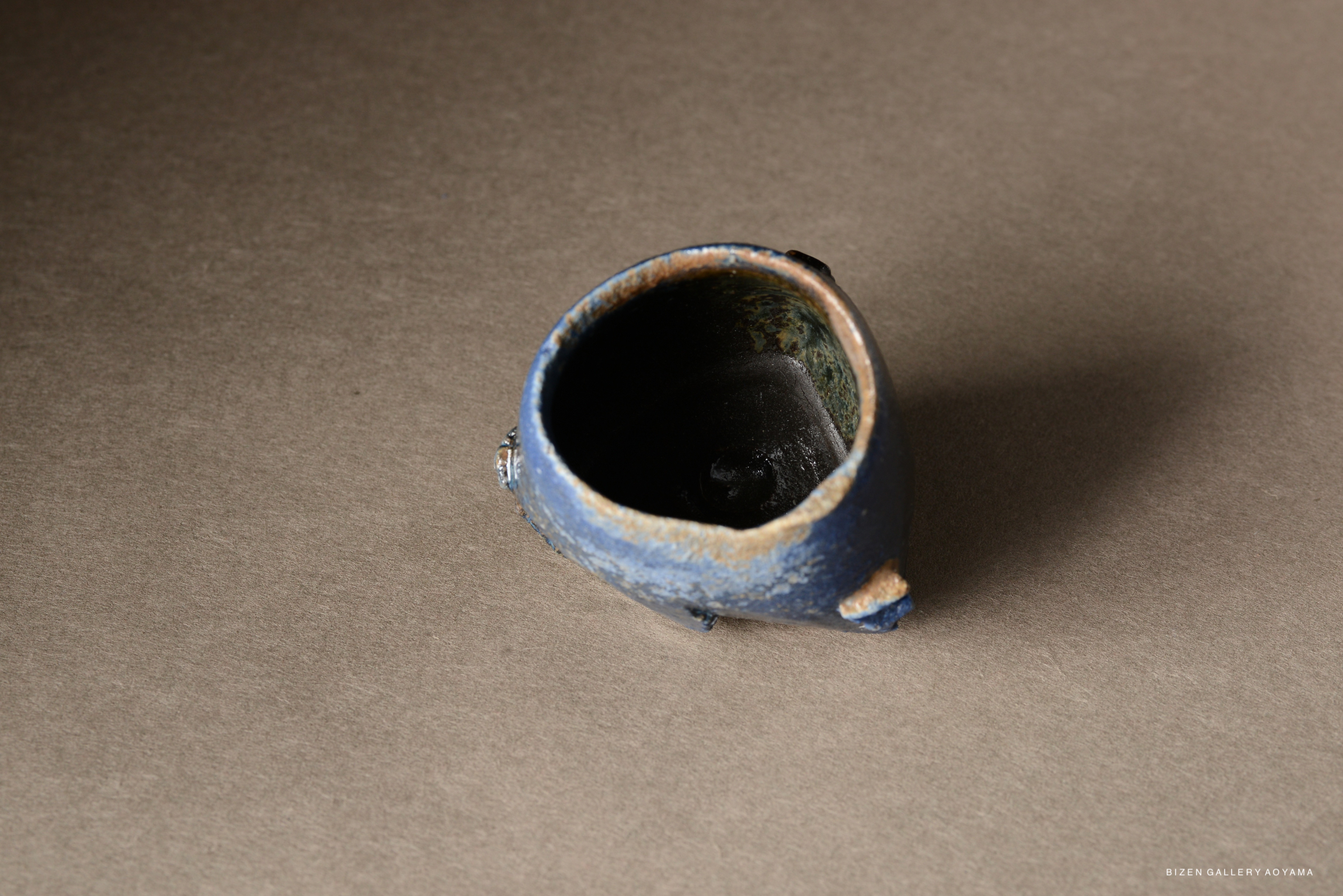 A small, irregularly shaped blue pottery piece with a dark interior, viewed from above against a gray background.