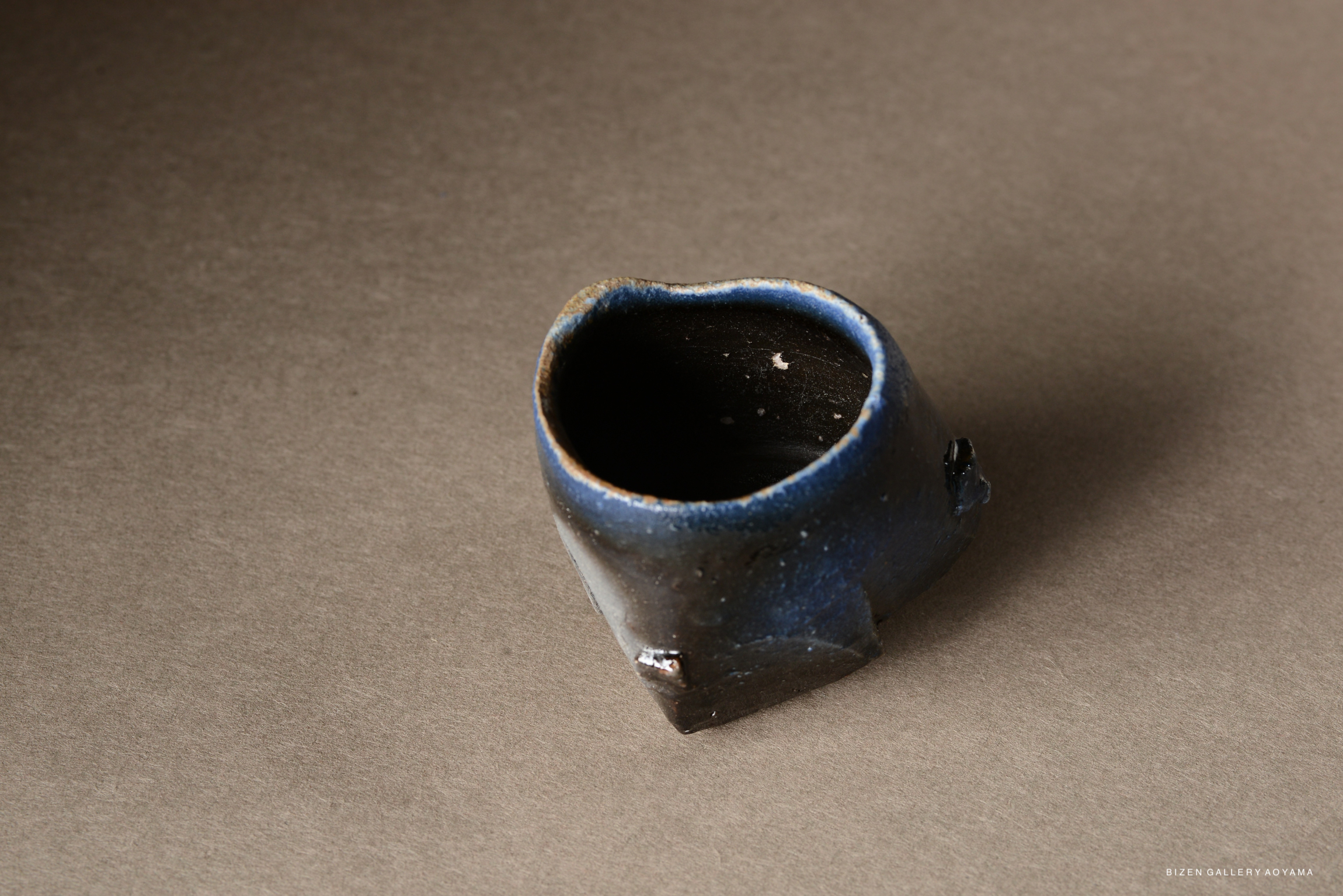 A small, unique ceramic cup with a heart-shaped opening and a dark blue glaze, resting on a textured brown surface.