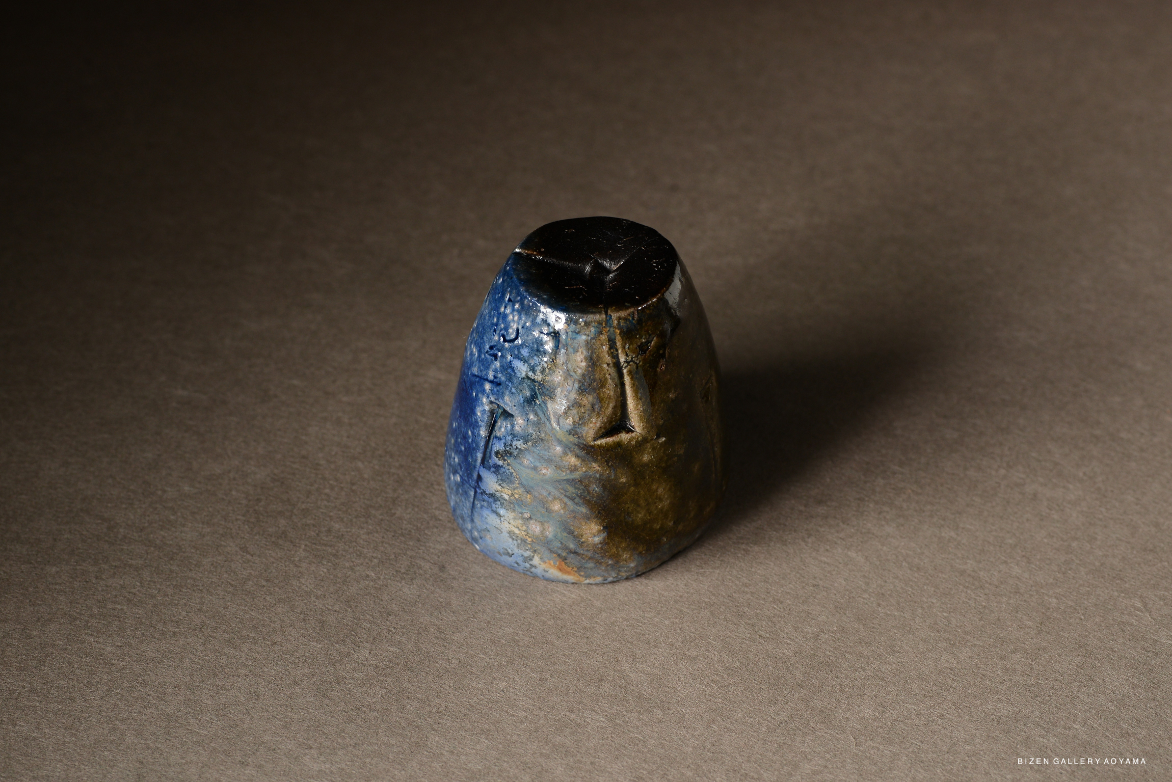 A small ceramic piece with a blue and brown glaze finish, shaped like a rounded vessel, sitting on a neutral background.