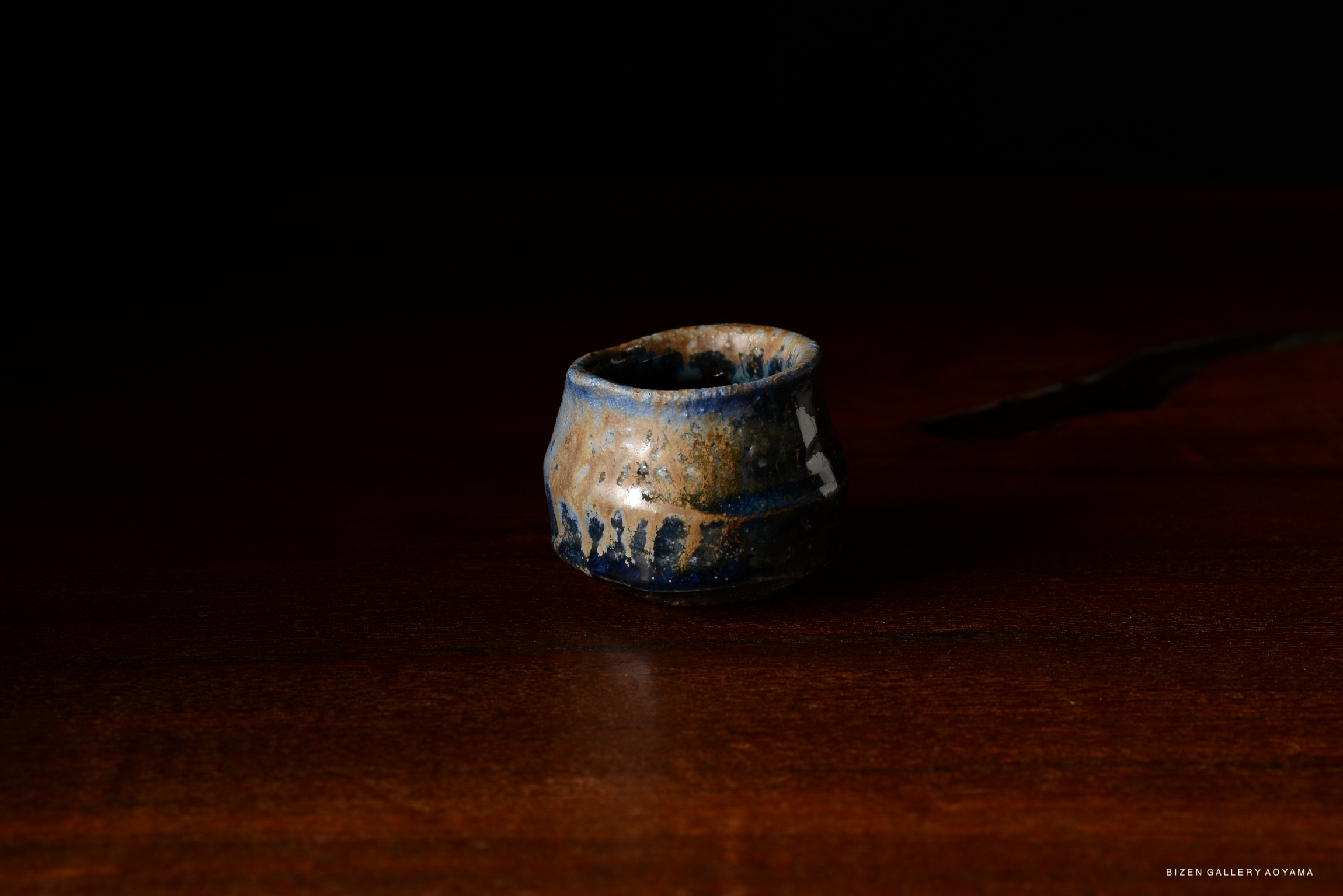 A handmade ceramic cup with a textured surface and blue and brown glaze, set against a dark background.