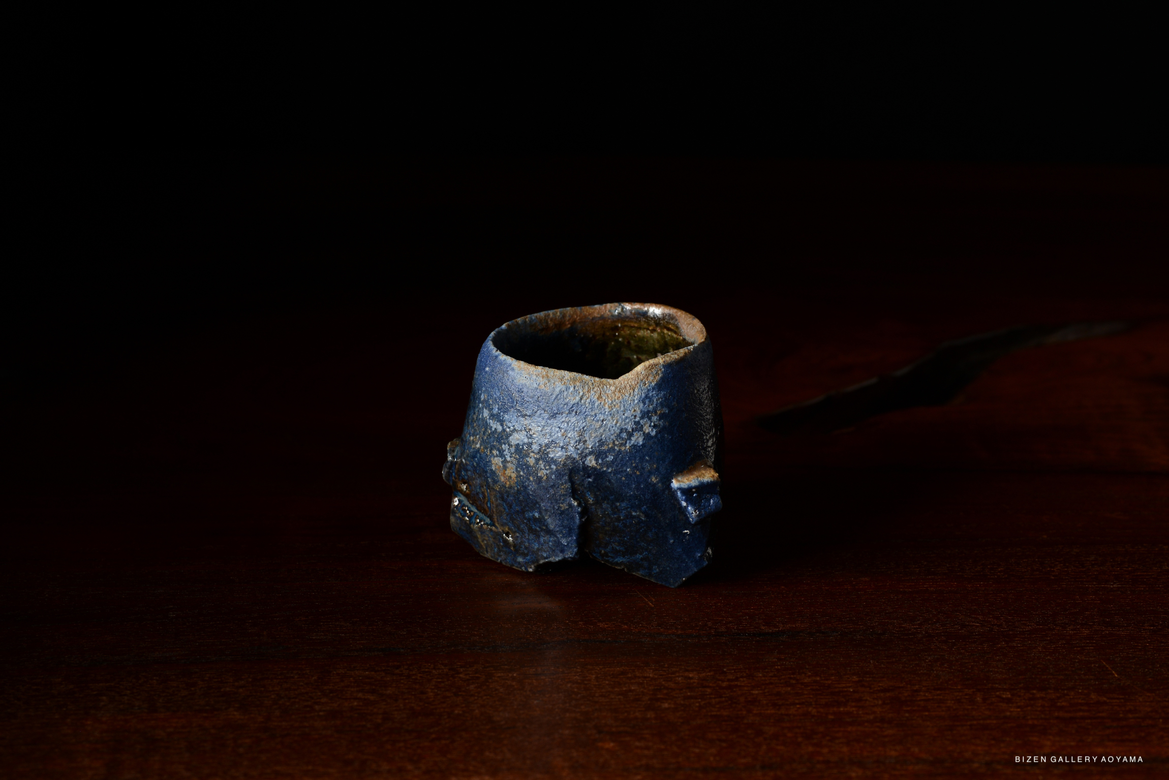 A close-up of a dark, textured tokkuri (sake vessel) sitting on a wooden surface against a black background.