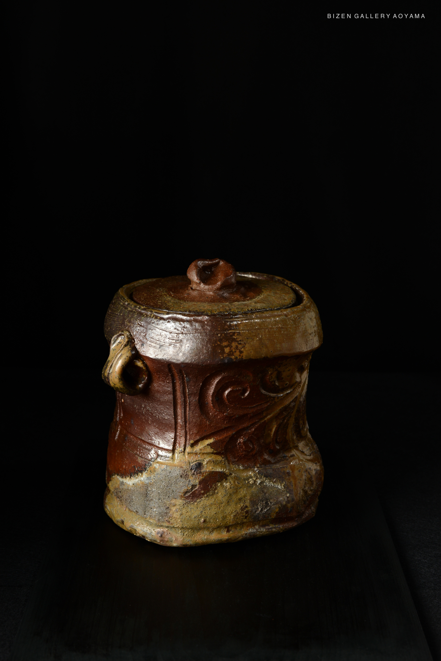 A Bizen mizusashi (water container) with intricate textures and earthy tones, featuring a curved shape and decorative carvings, set against a dark background.