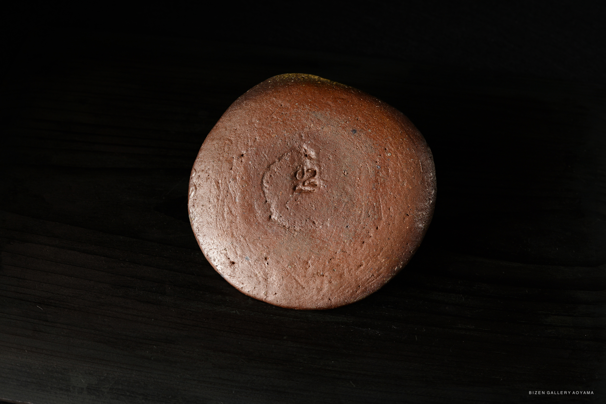 A top view of a Bizen Mizusashi ceramic piece, featuring a smooth, reddish-brown surface with a subtle texture and markings, resting on a dark wooden surface.