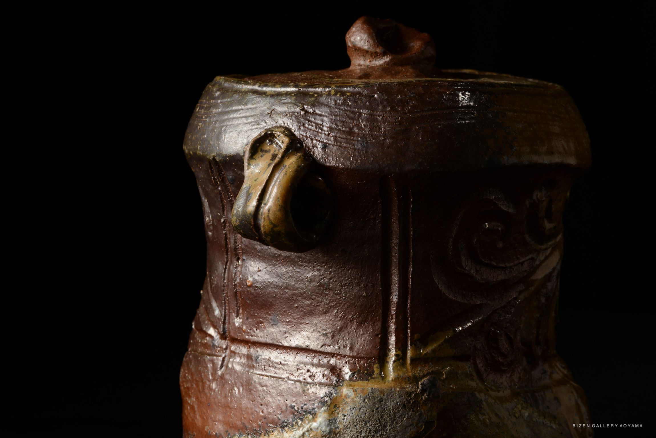 Close-up of a Bizen Mizusashi teapot with intricate patterns, featuring a round lid and handle, displayed against a dark background.