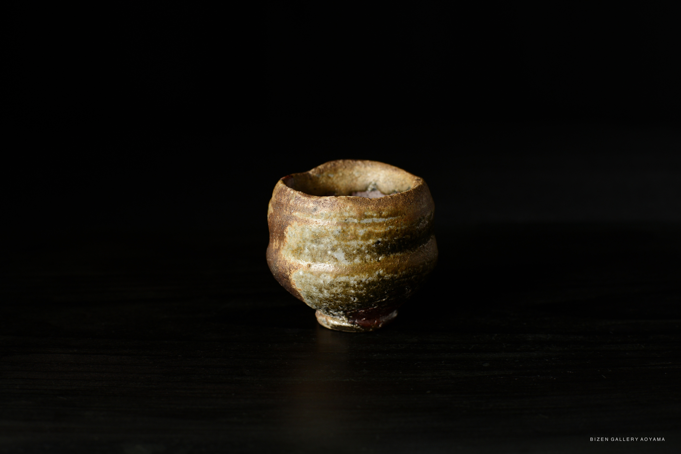 A close-up view of a traditional Bizen pottery Guinomi sake cup, showcasing its unique texture and color against a dark background.