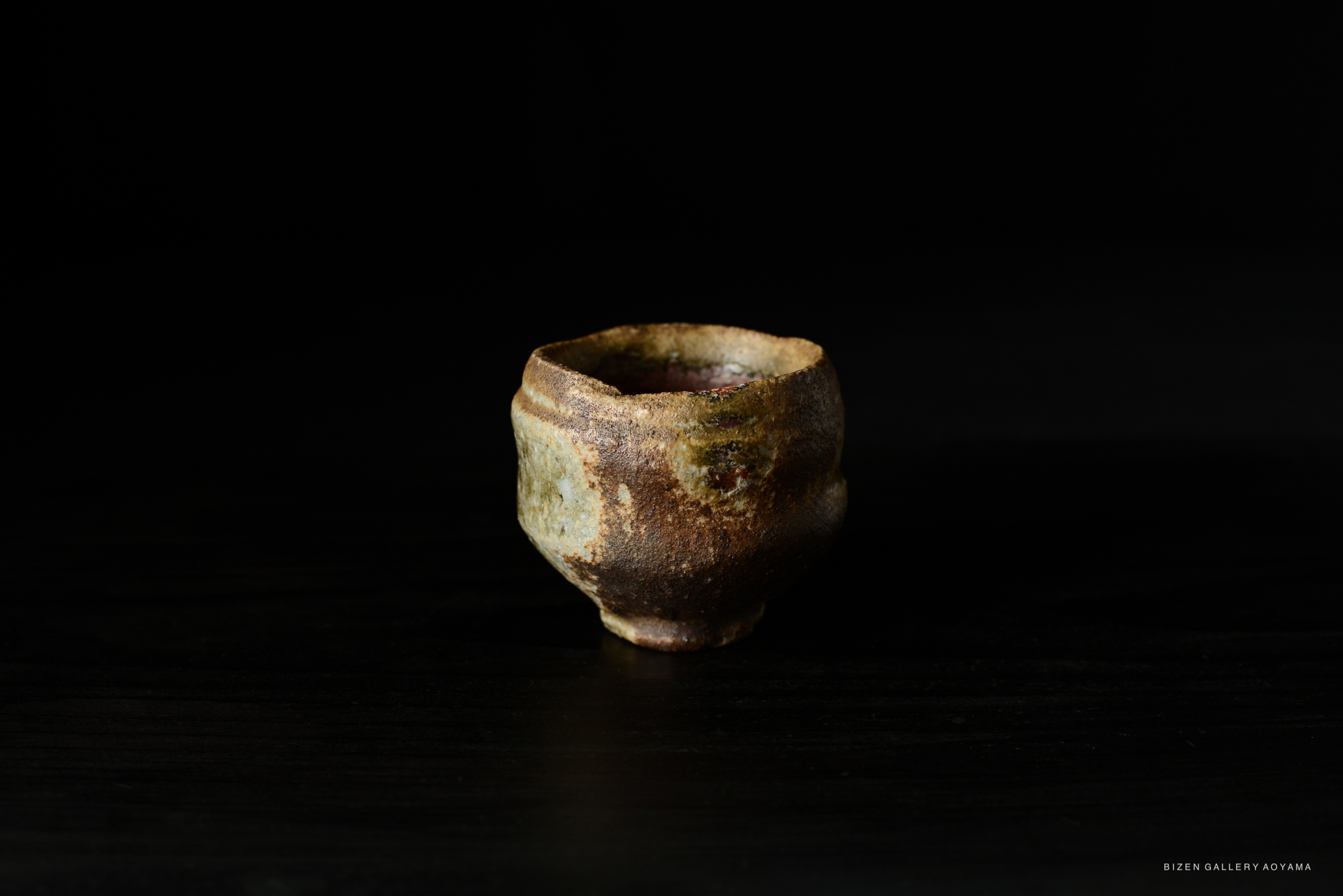 A handcrafted Bizen Guinomi sake cup with a rustic finish, sitting on a dark surface against a black background.