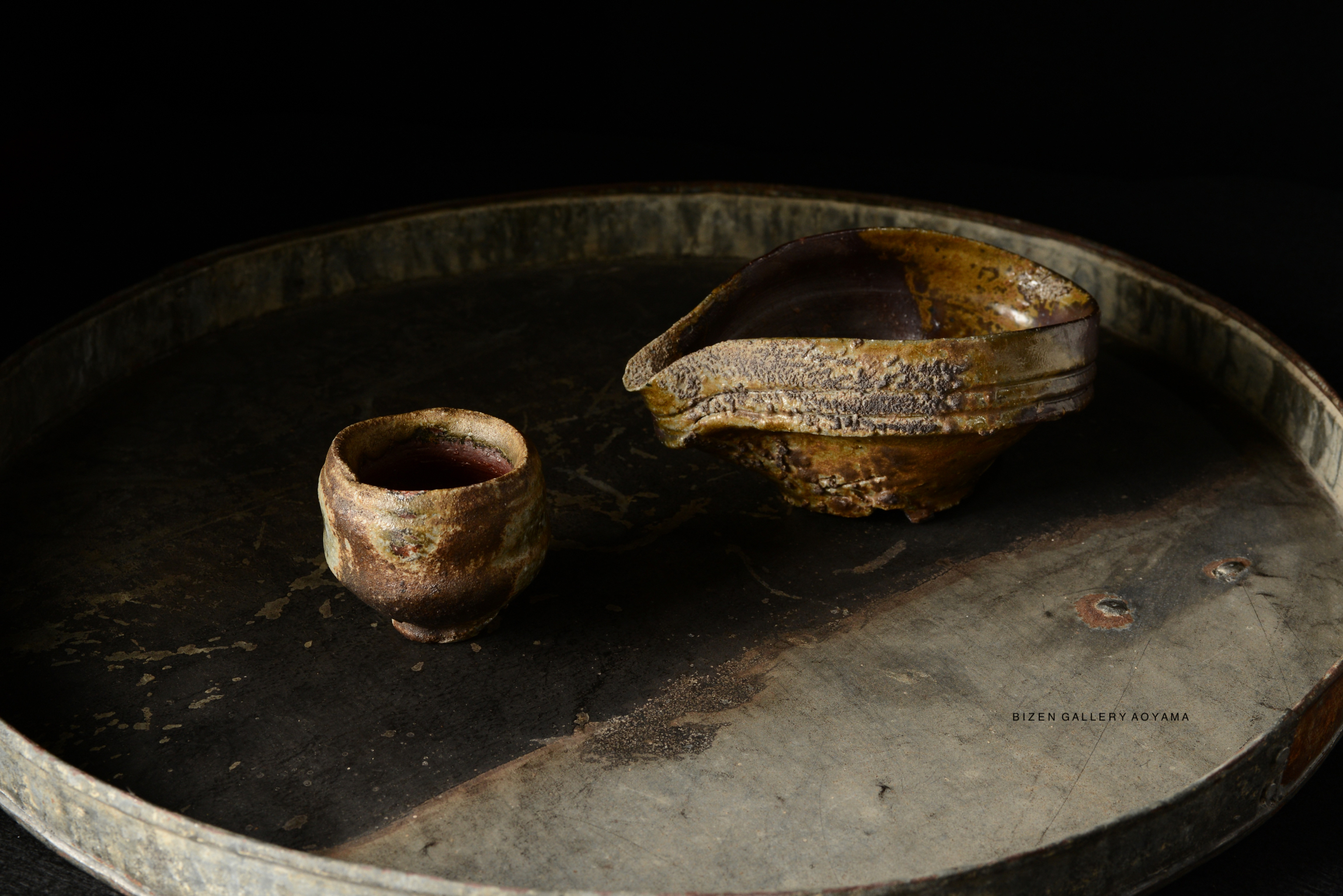 A traditional Bizen pottery tea cup and a visually unique serving bowl on a round, weathered metal tray against a dark background.