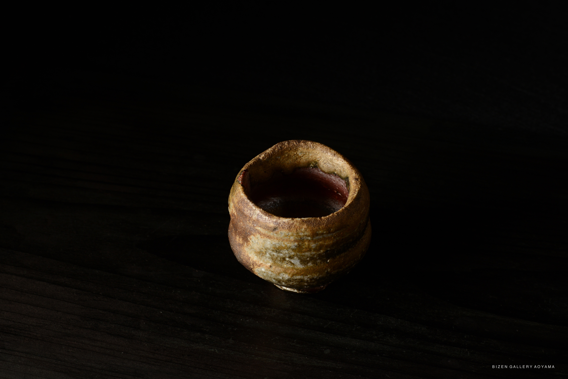 A close-up of a Bizen Guinomi sake cup, showcasing its earthy tones and textured surface against a dark background.