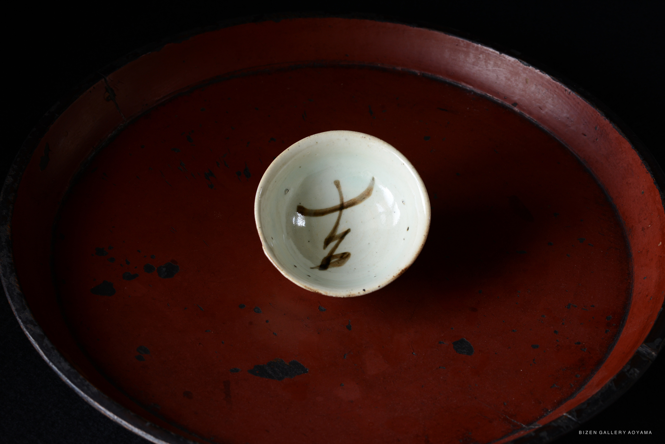 A small, pale ceramic sake cup with a simple calligraphic design on the interior, placed on a dark red lacquer tray.