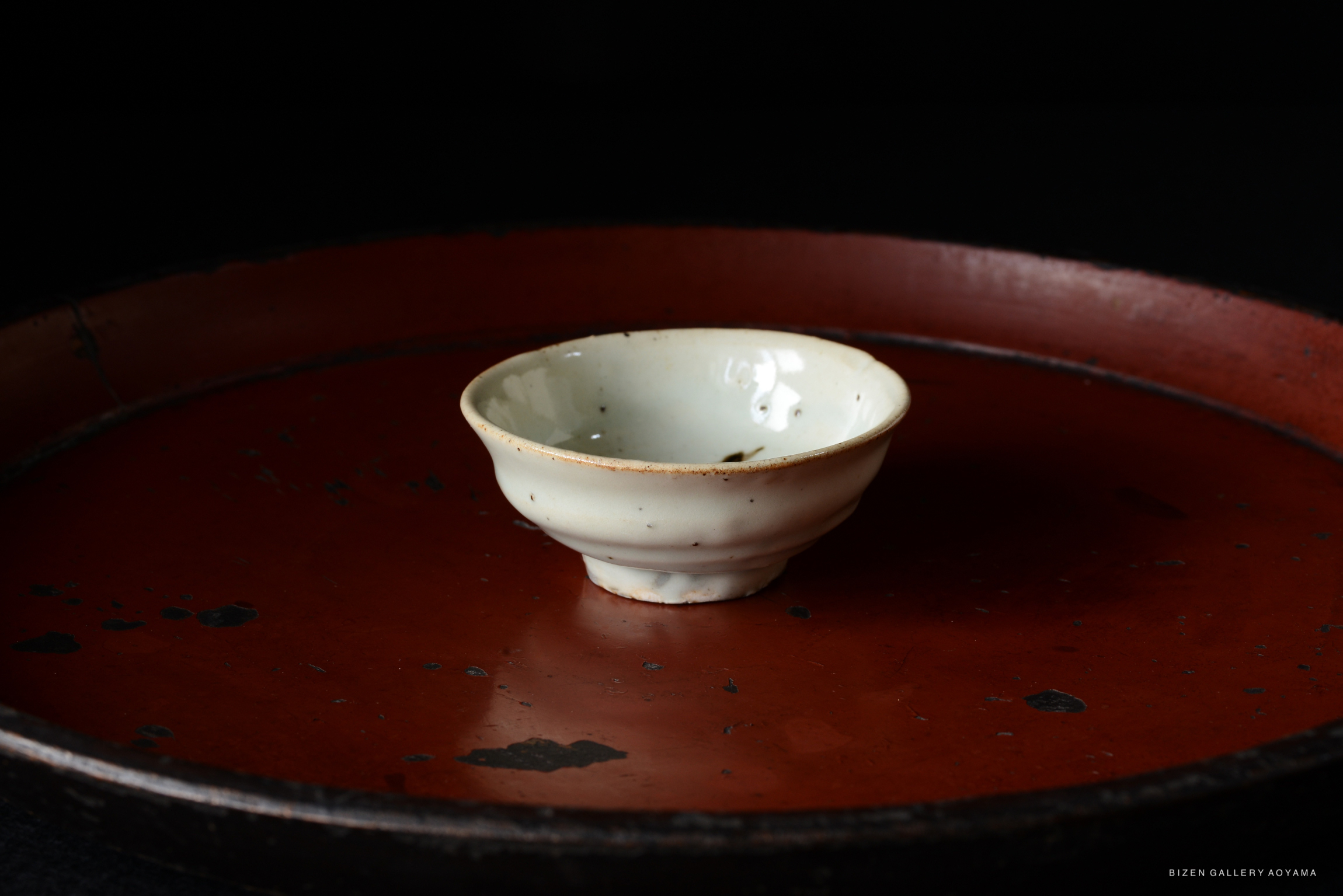 A small, white ceramic bowl with a subtle glaze sits on a large, round, reddish-brown tray against a dark background.