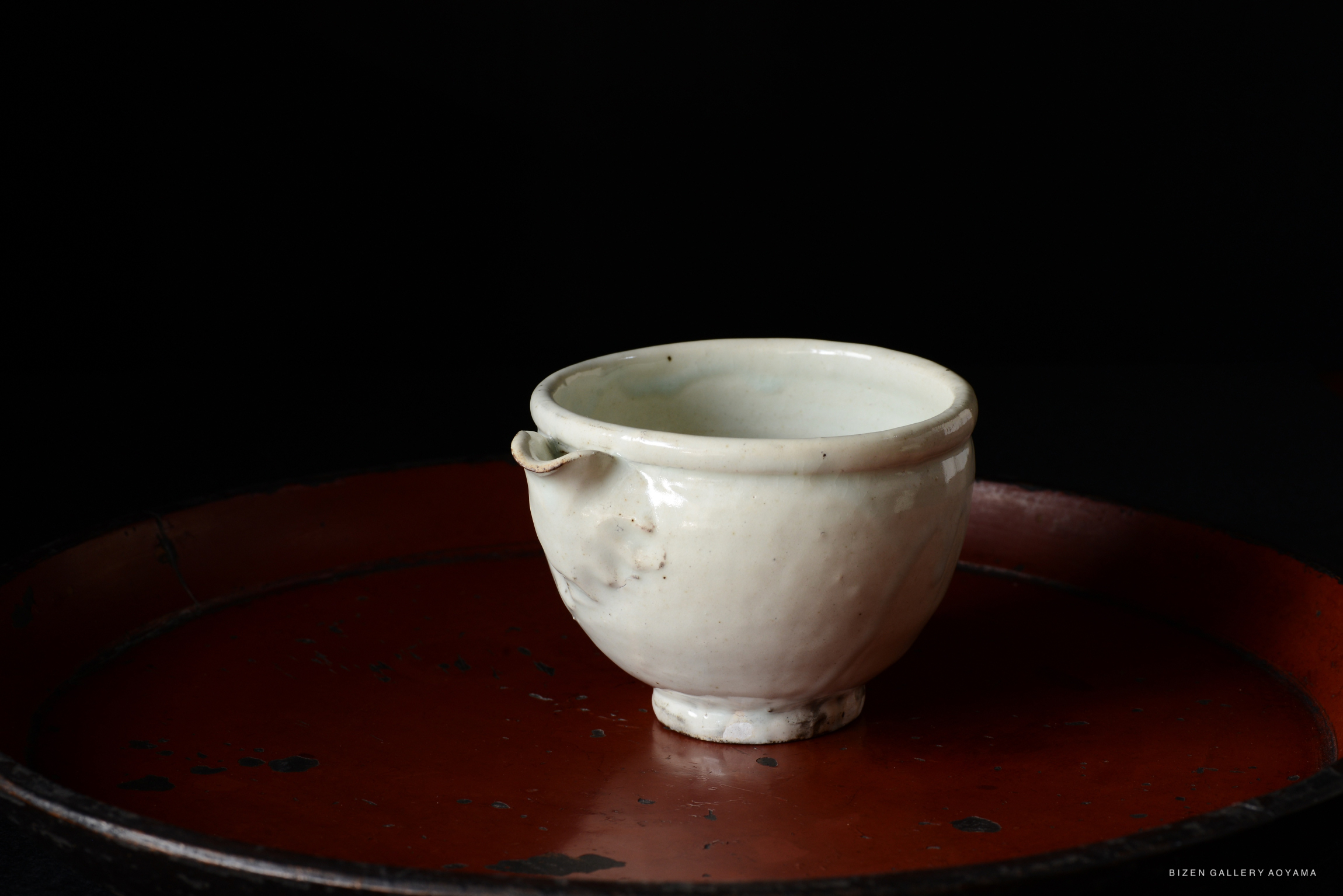 A white ceramic cup with a small spout, resting on a red circular tray against a dark background.