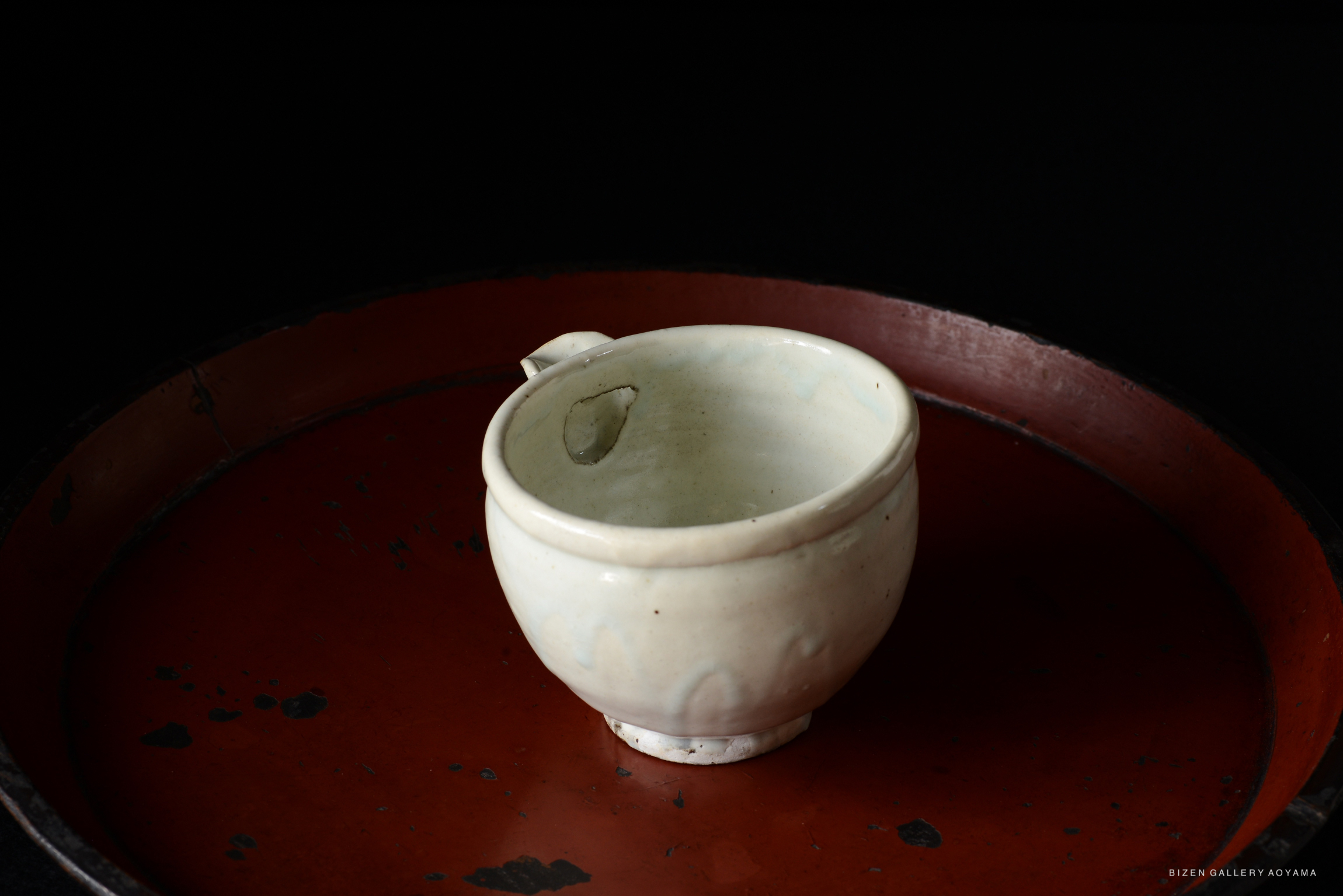 A handmade ceramic cup displayed on a round red tray against a dark background.