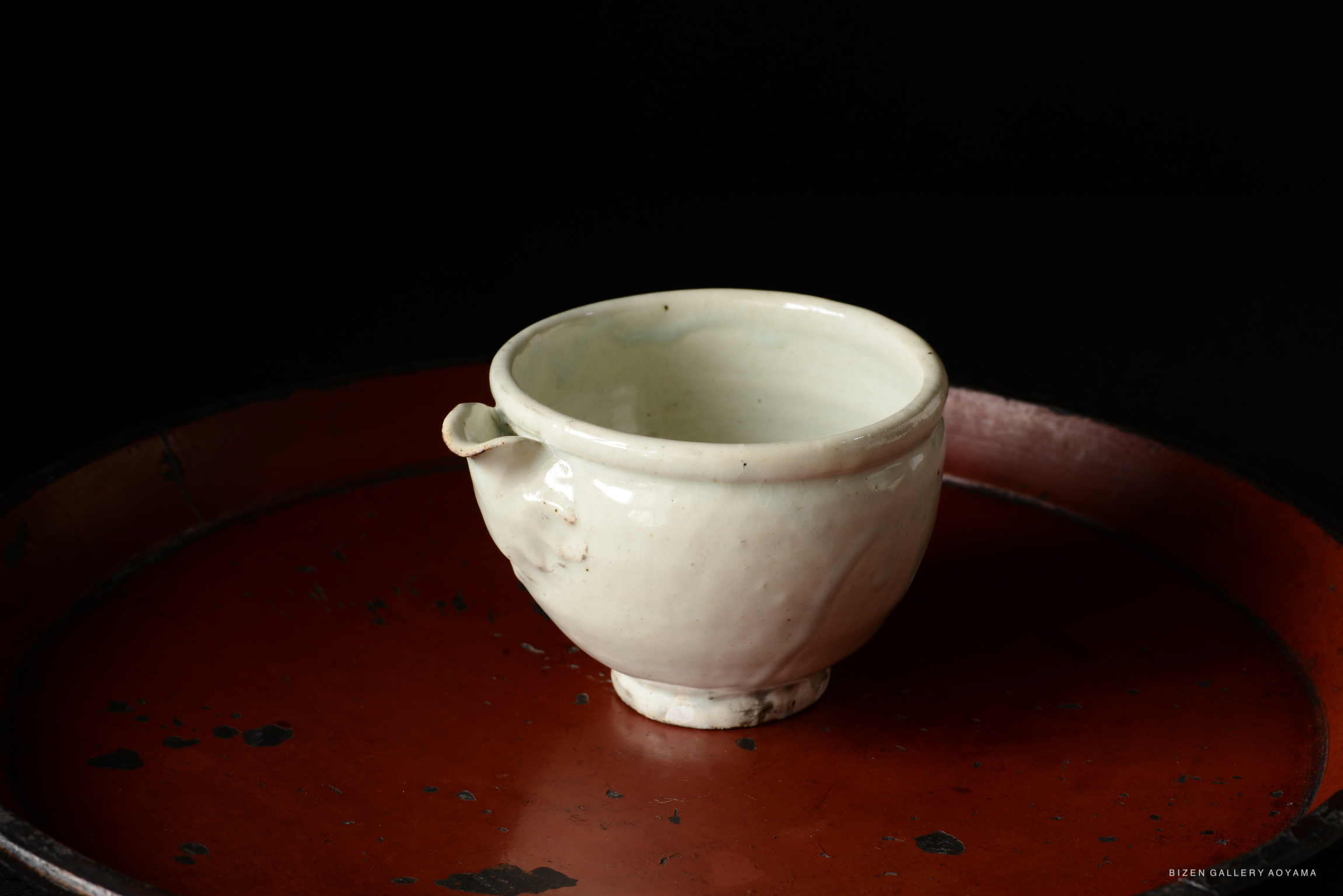 A handcrafted white ceramic cup with a small handle, placed on a red lacquer tray against a black background.