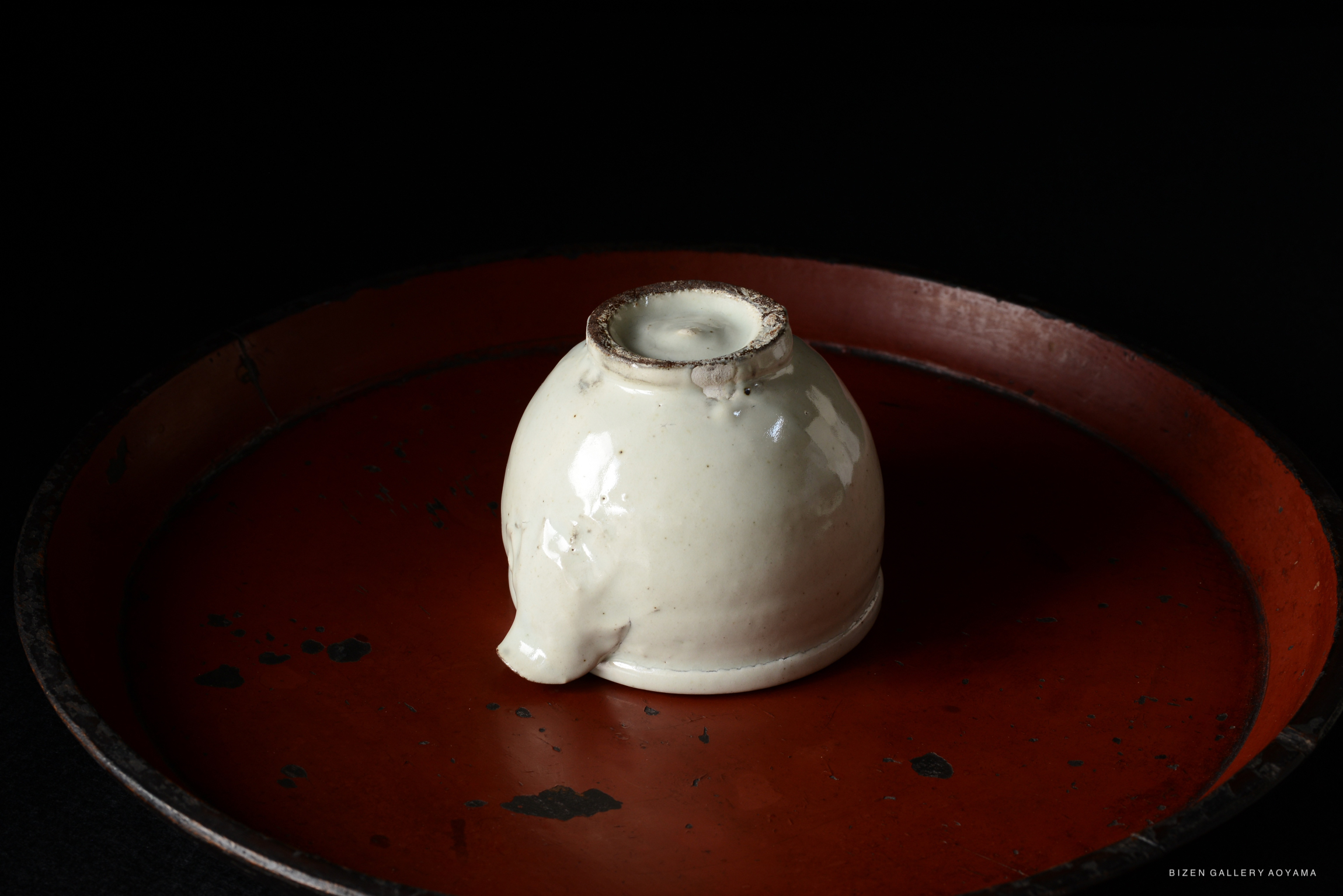 A white ceramic vessel with a curved shape resting on a round, slightly tarnished red tray, against a black background.