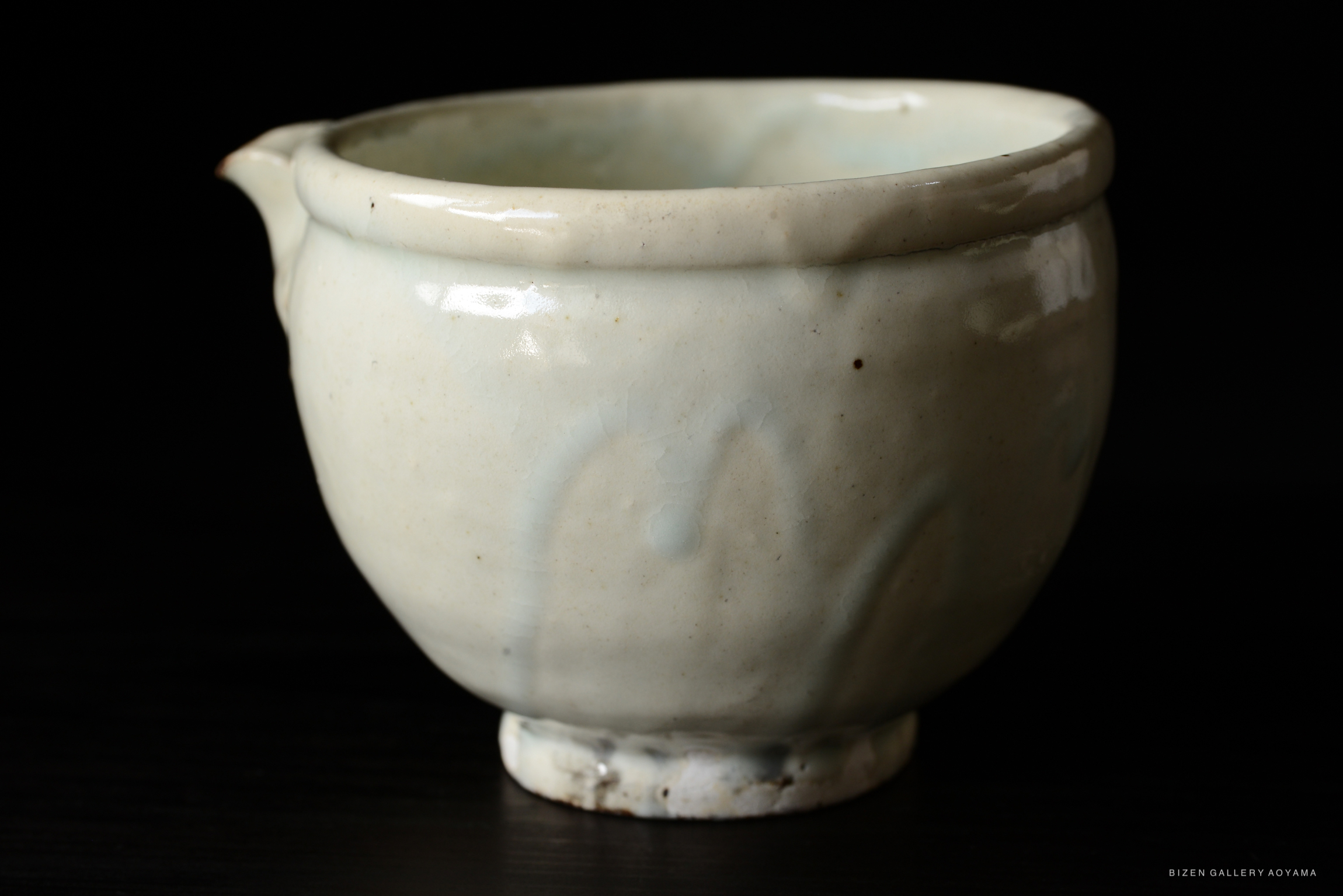 A close-up of a light-colored ceramic cup with a curved spout and subtle design patterns, set against a dark background.