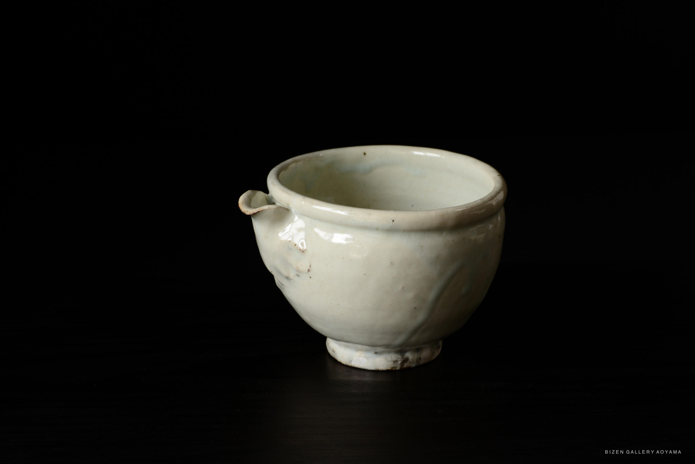 A small, white ceramic bowl with a spout, featuring a smooth surface and a round foot, set against a dark background.