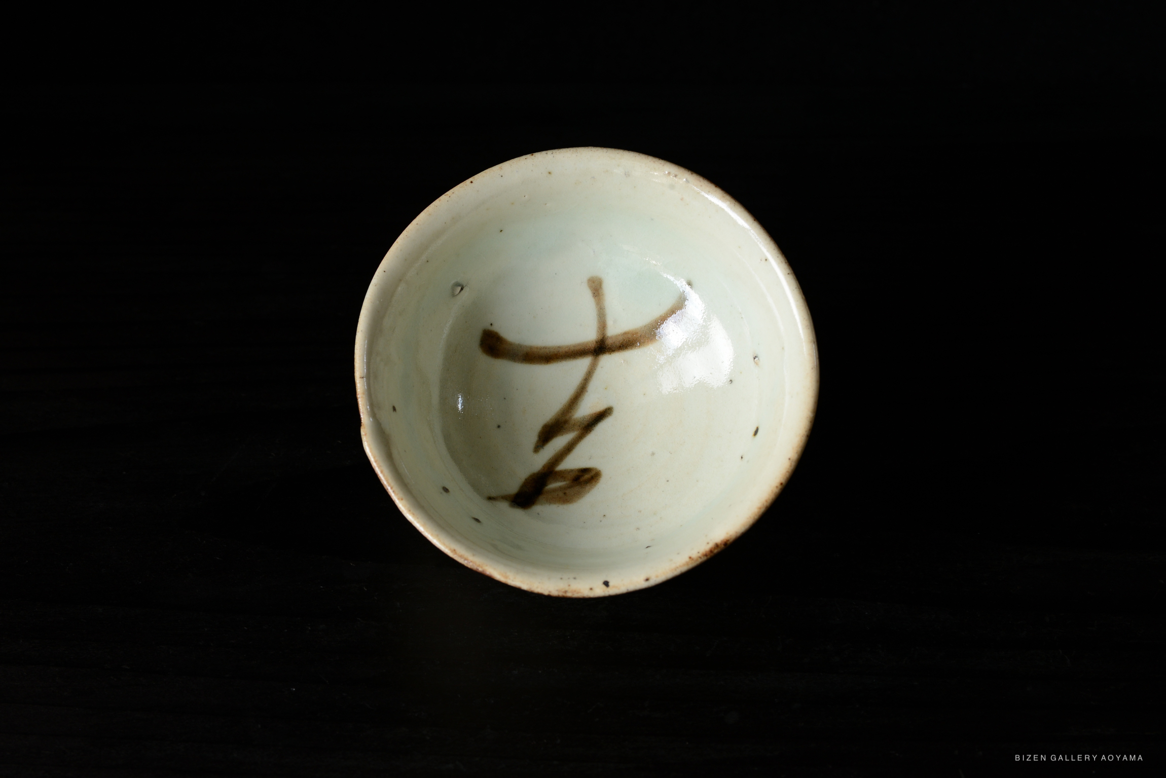 A close-up view of a pale green ceramic bowl featuring a dark brown calligraphic design inside.