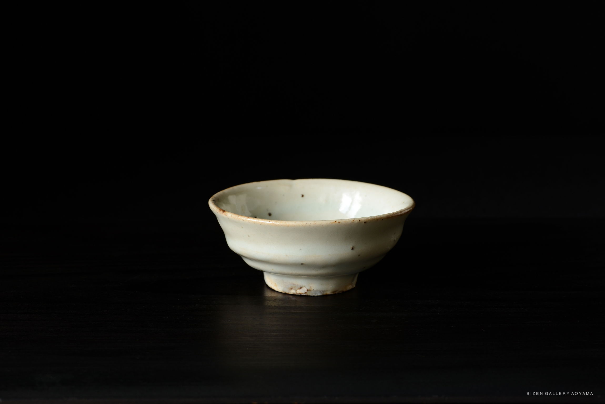 A small, delicate ceramic bowl with a light glaze, featuring subtle imperfections and a slightly flared rim, set against a dark background.