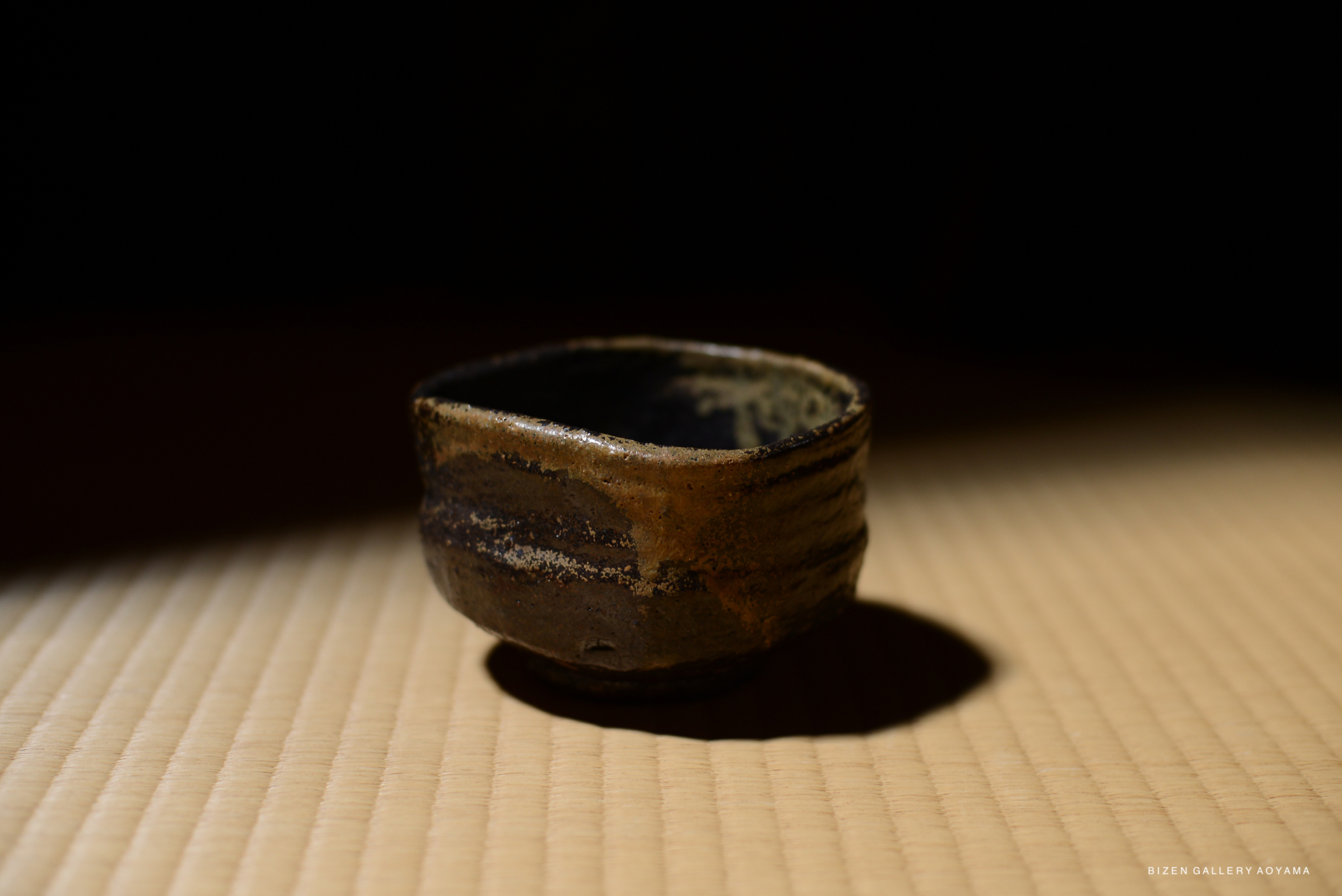 A small, rustic ceramic tea bowl placed on a tatami mat, with a focus on its textured surface and natural glaze.