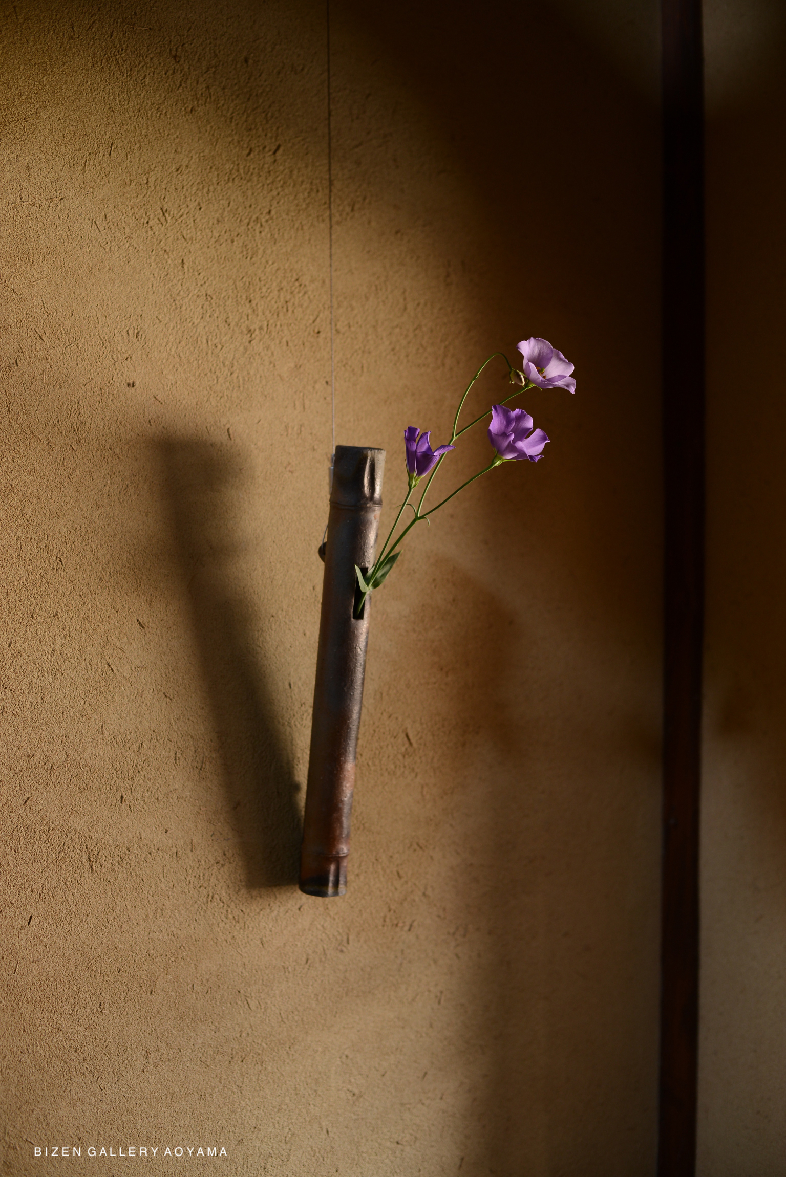 A bamboo vase with purple flowers hanging against a textured beige wall.