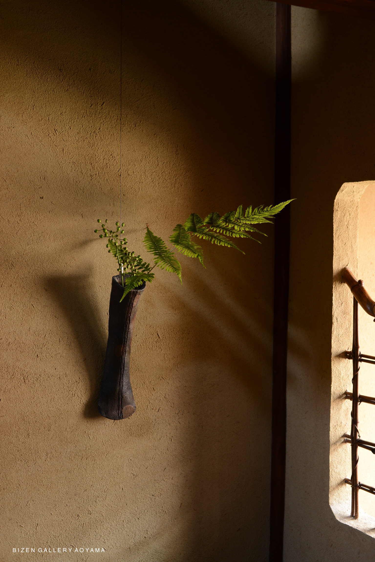 A wall-mounted vase holding green ferns against a textured beige wall.