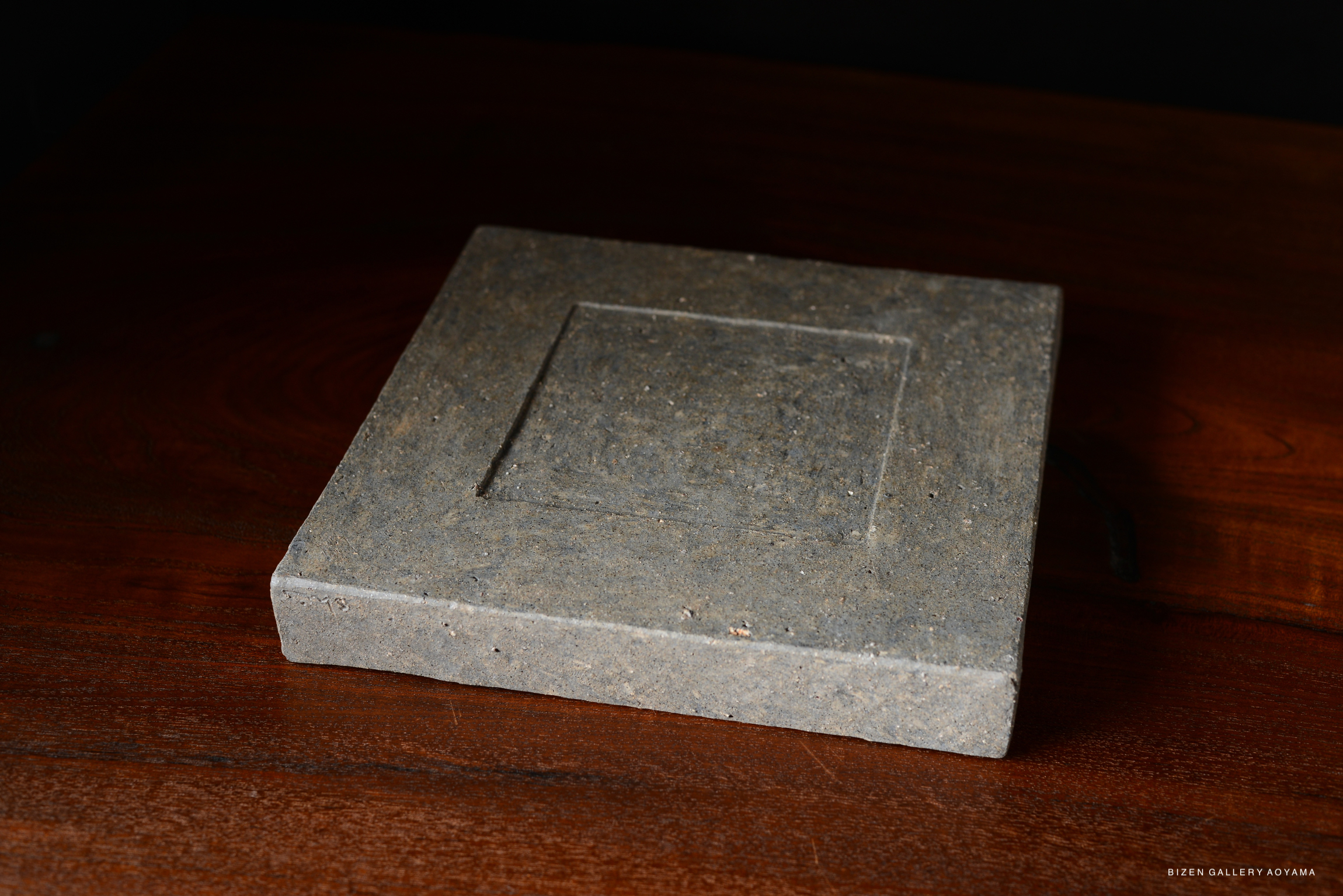 A square plate made of dark stone, featuring a smooth surface and a simple, modern design, placed on a wooden table.