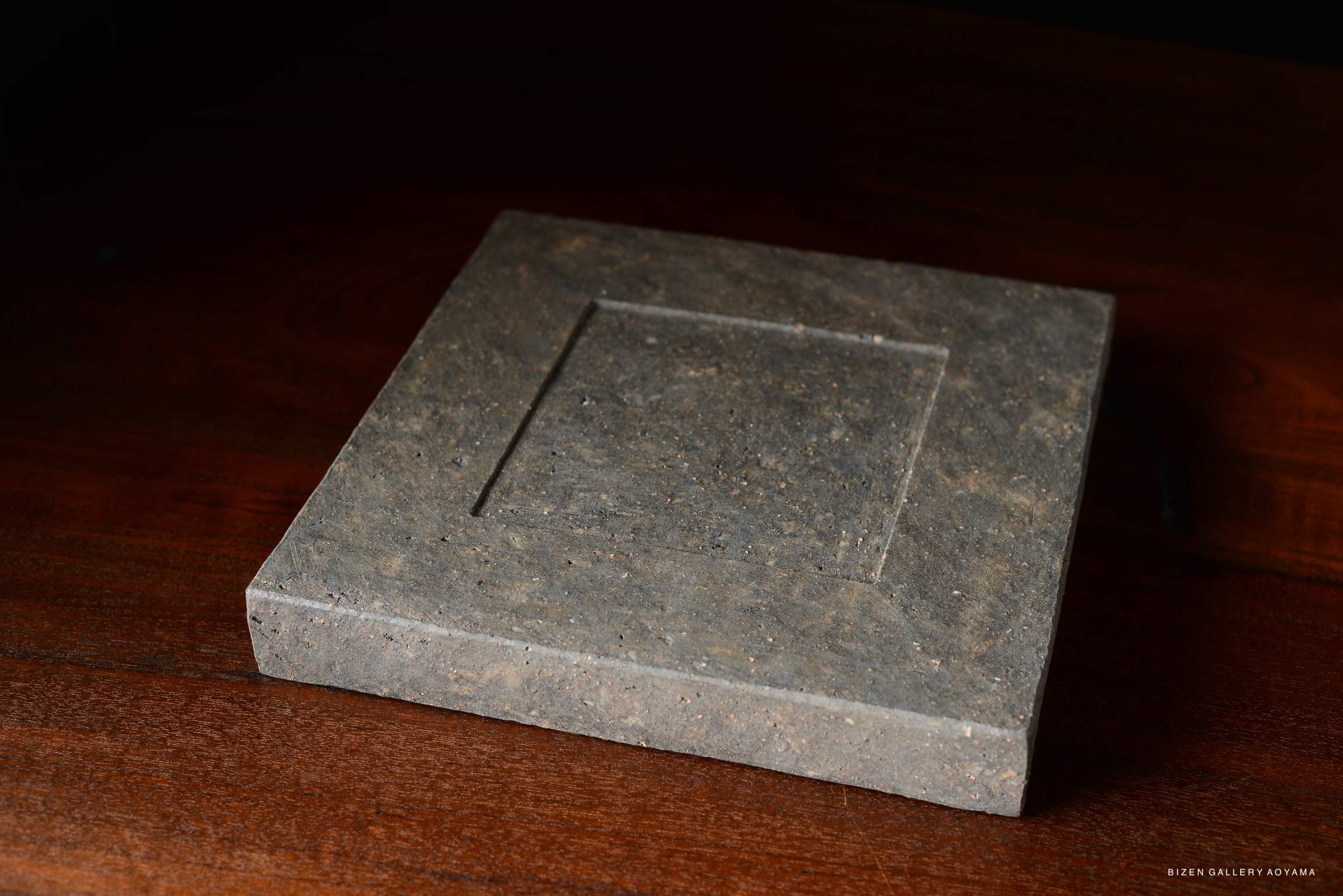 A square plate made of grey pottery, featuring a subtle textured surface, placed on a wooden table.
