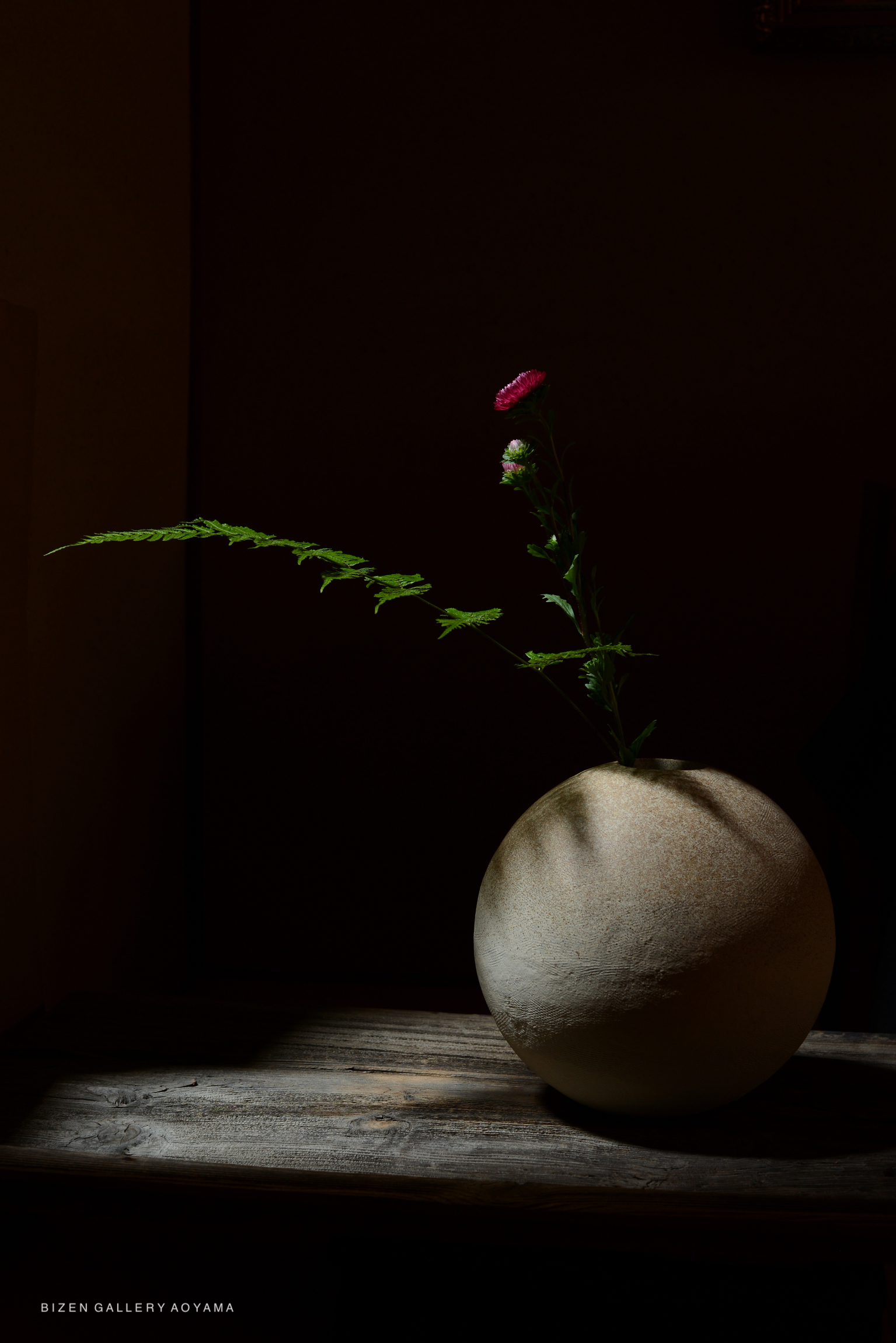 A round Bizen pottery vase with a pink and green flower arrangement on a wooden surface illuminated by soft lighting.