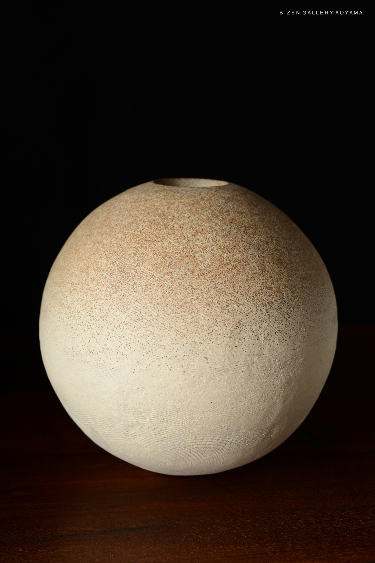 A round, beige ceramic vase with a textured finish, resting on a wooden surface against a dark background.