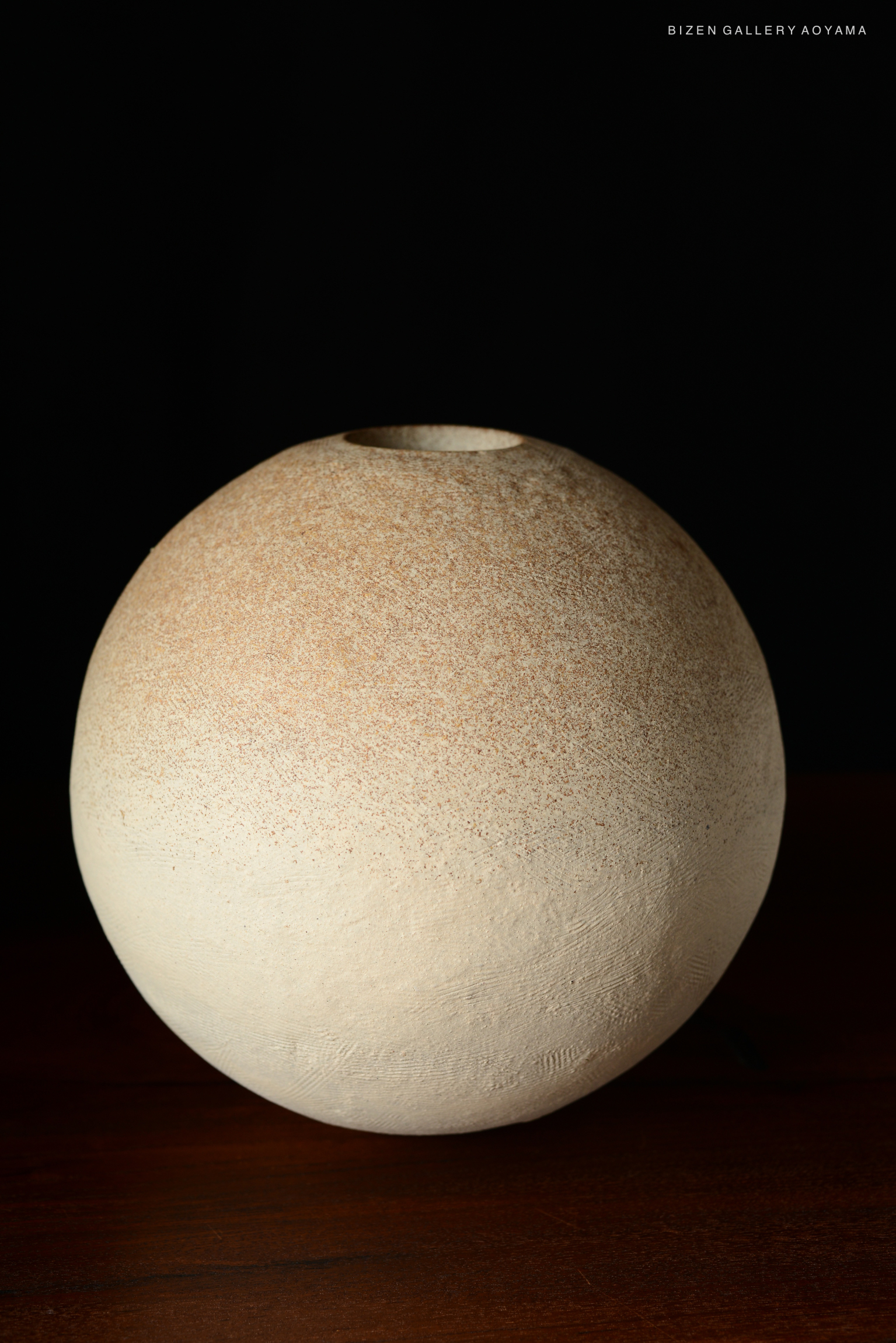 A round beige ceramic vase with a textured surface, showcasing a subtle gradient from light to darker shades, placed on a wooden surface against a black background.
