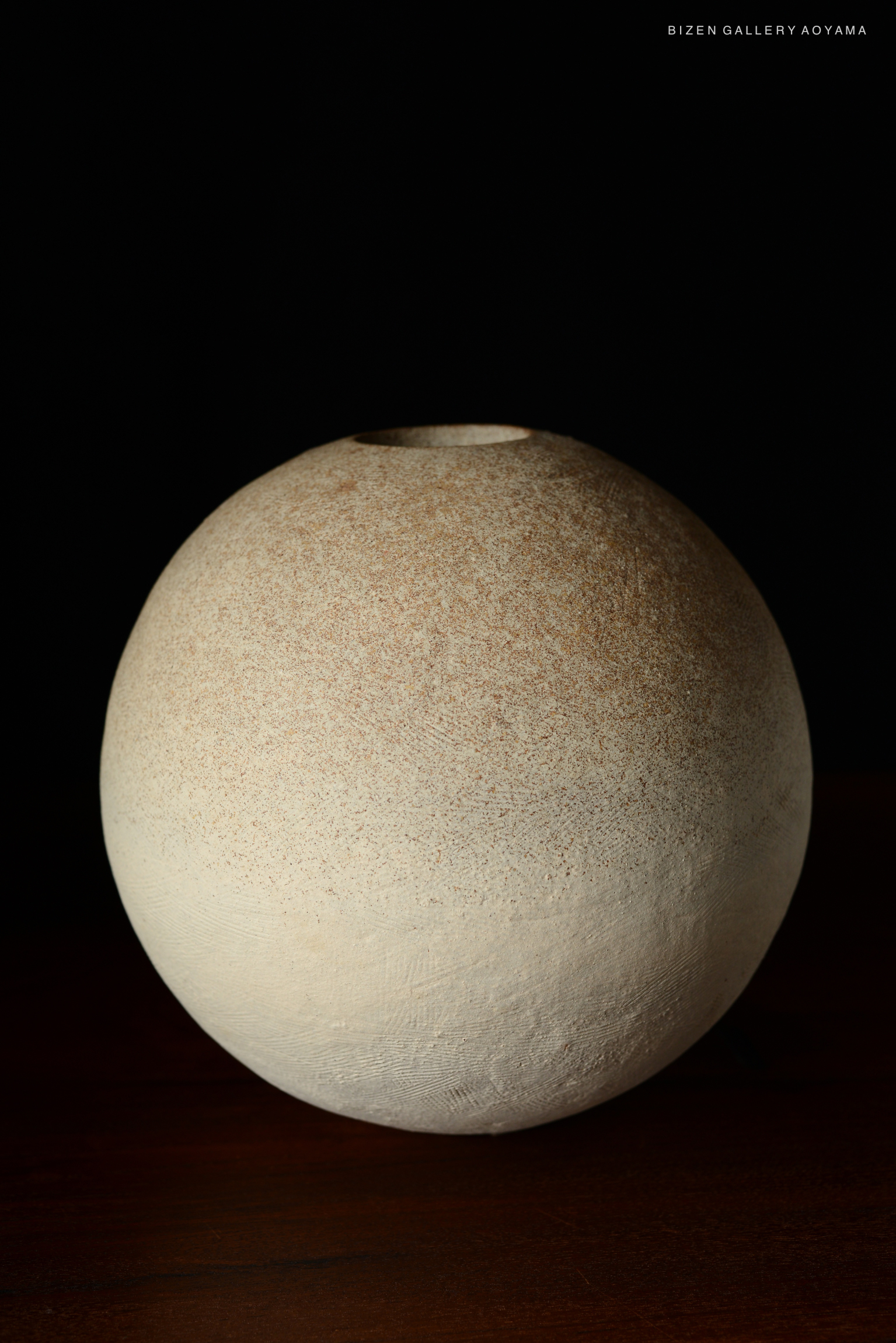 A round, minimalist Bizen vase with a textured surface, showcasing a subtle beige color against a dark background.